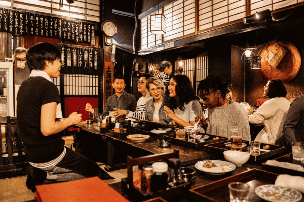 A group of friends enjoying a lively, casual dining experience inside a traditional Japanese Izakaya, sitting at a low wooden table with small plates and drinks.