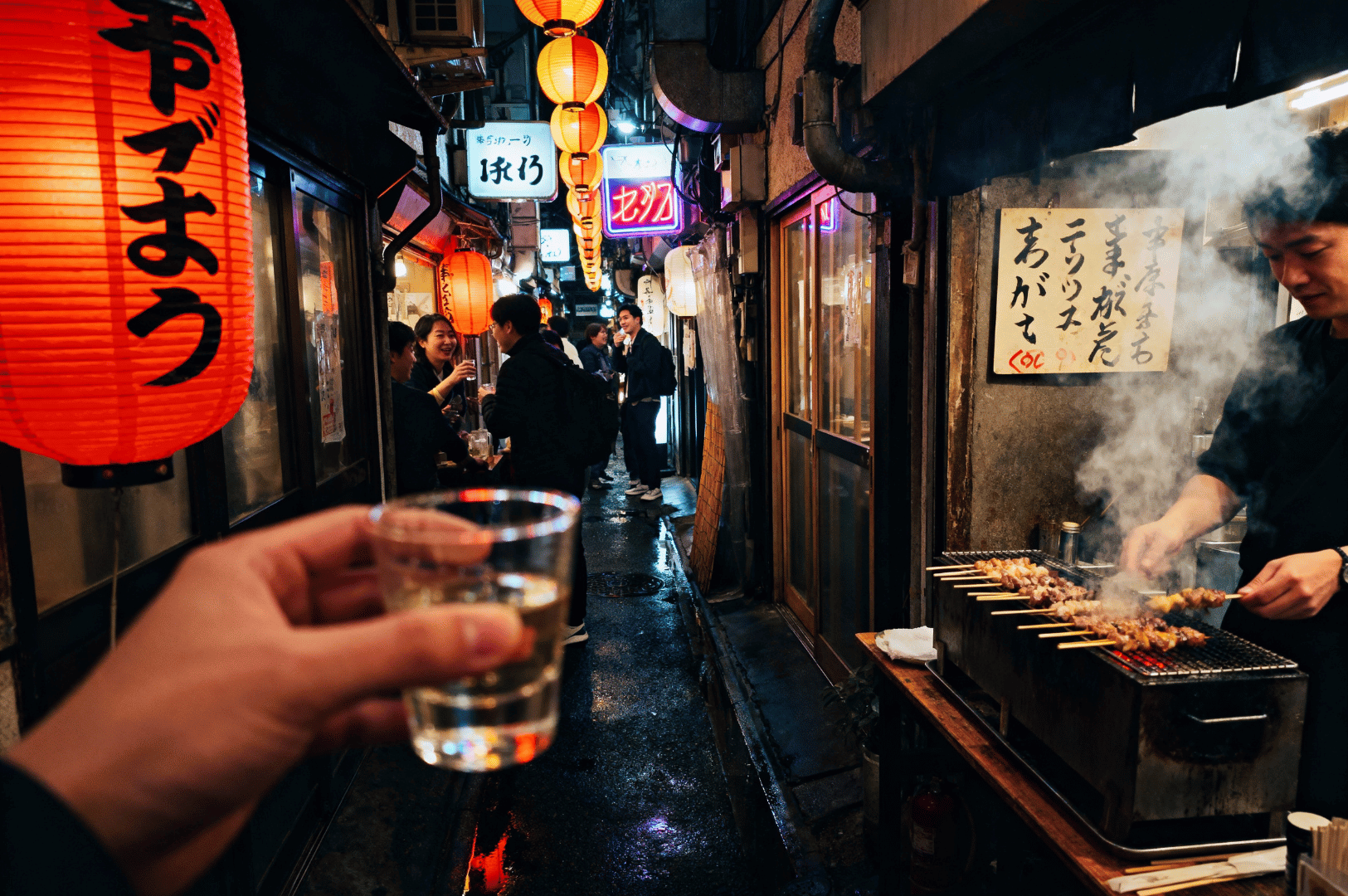 Hand holding a glass of sake in a lantern-lit alleyway lined with izakayas and street grills—capturing Japan’s after-work dining culture and entrance etiquette in traditional pubs.