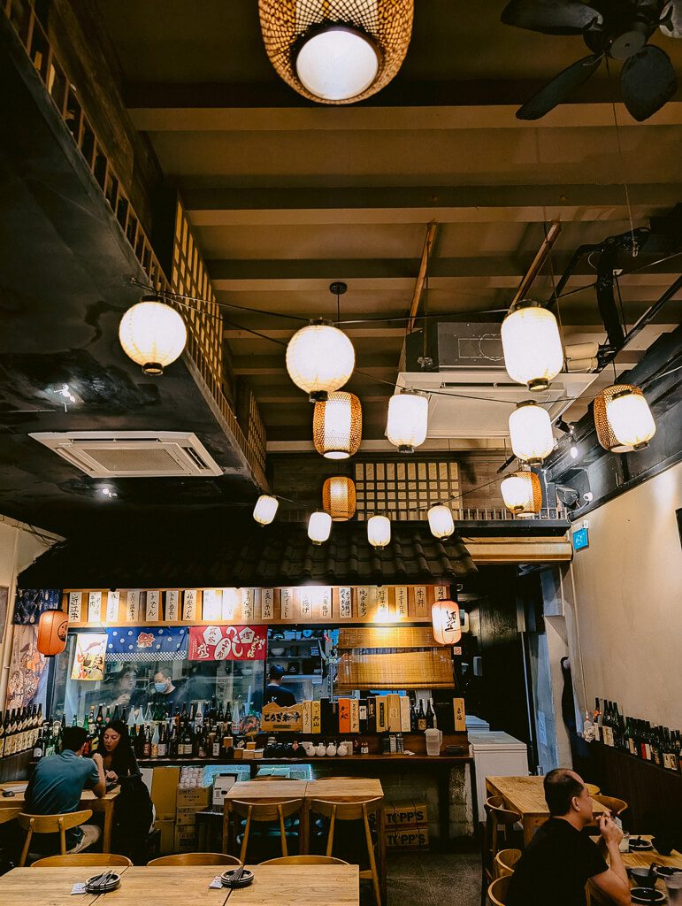 Cozy, authentic Izakaya restaurant interior with hanging bamboo-style lanterns, low wooden tables, and a bar counter lined with sake bottles, capturing the traditional, casual Japanese dining atmosphere.