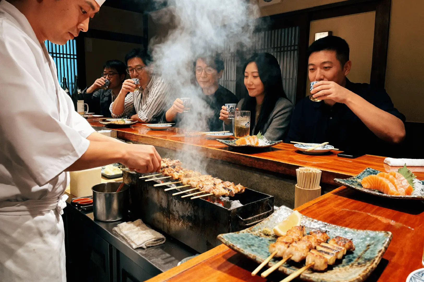 Chef grilling yakitori skewers over charcoal at an izakaya counter with customers dining and drinking in Singapore