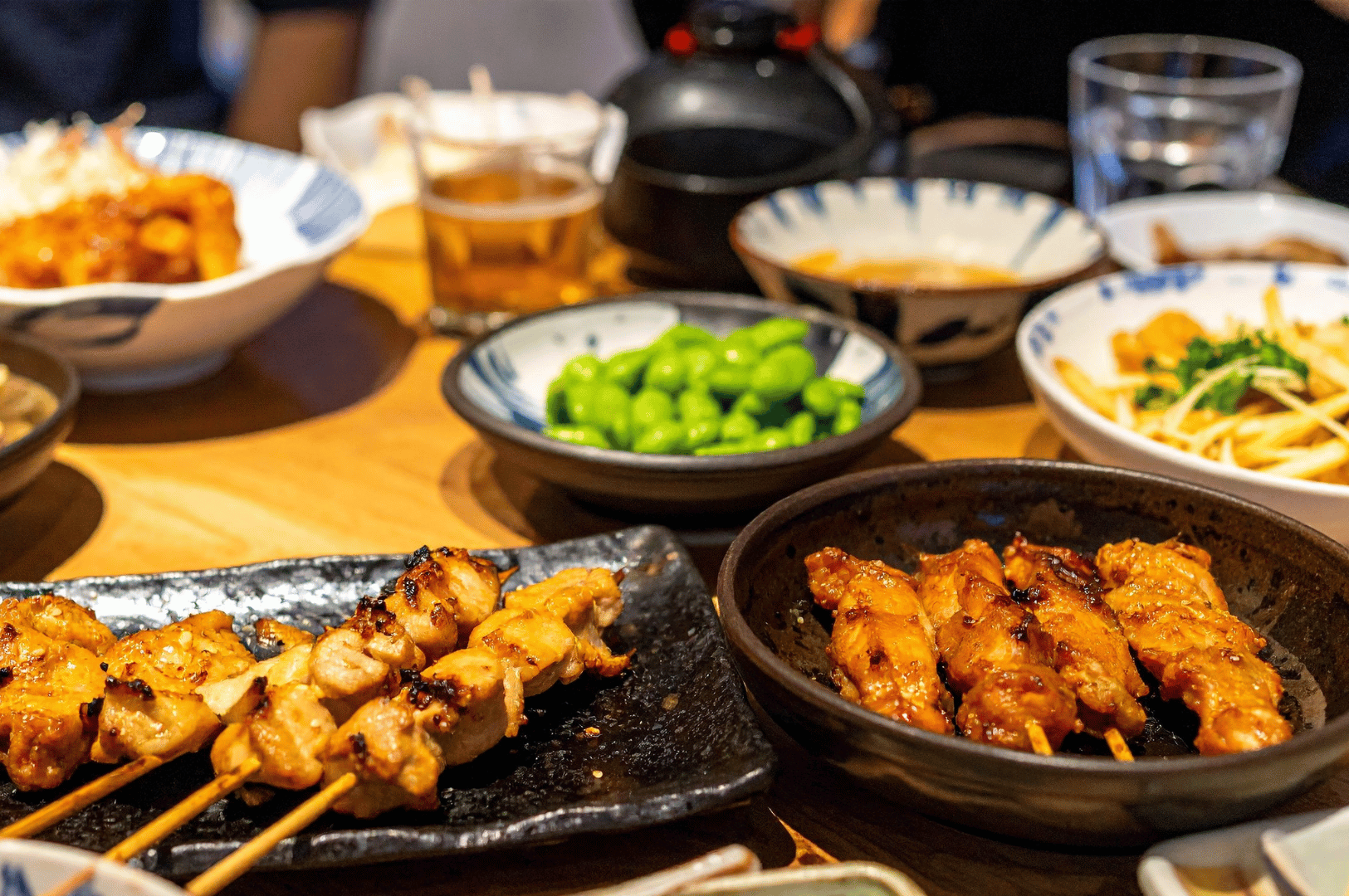 Grilled yakitori skewers and shared izakaya dishes on a wooden table, with edamame and small plates in a cozy restaurant setting in Singapore.