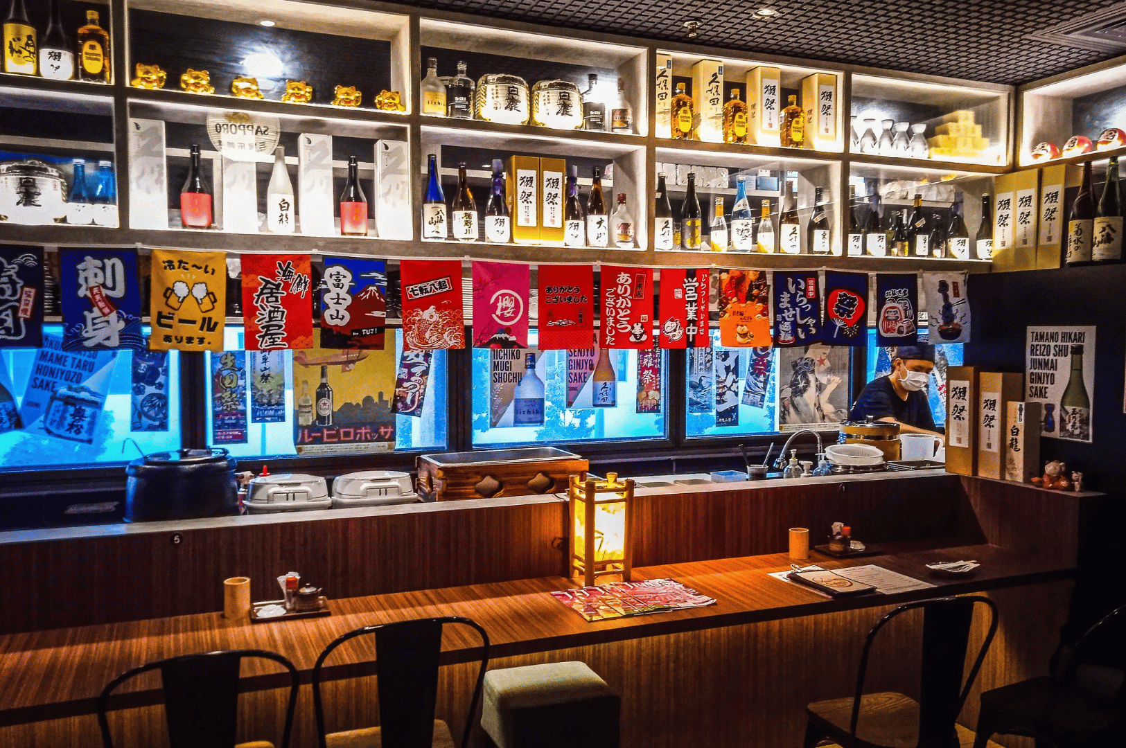 Cozy Izakaya Hikari bar with colorful Japanese banners, sake bottles on shelves, and a warm counter seating area in Singapore.