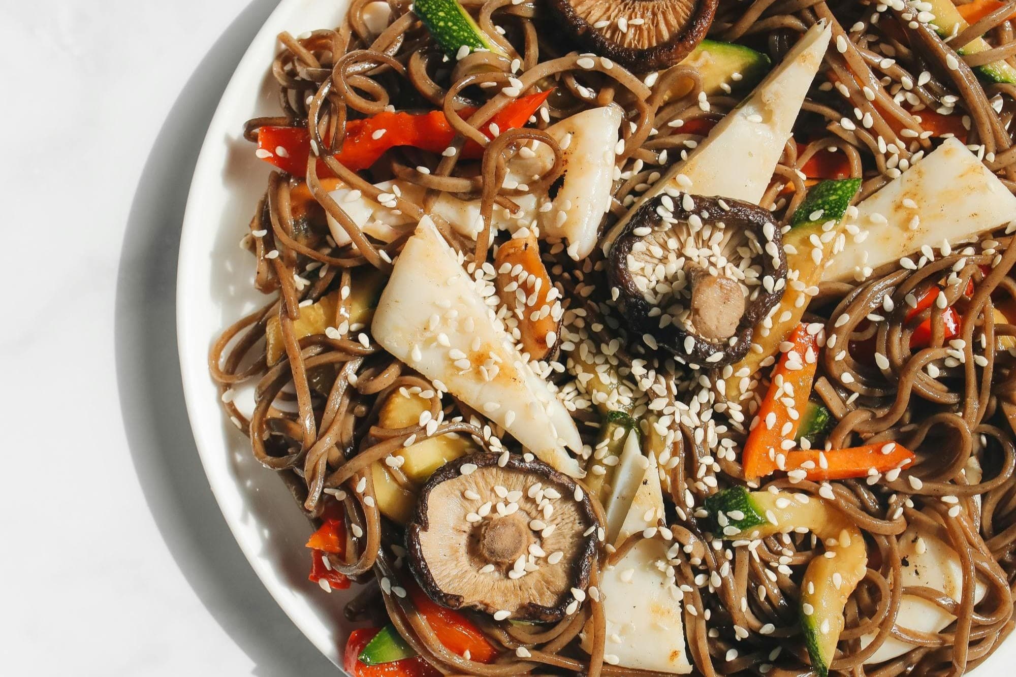 A close-up image of brown noodles beautifully decorated with various plant base foods like mushrooms and vegetables.