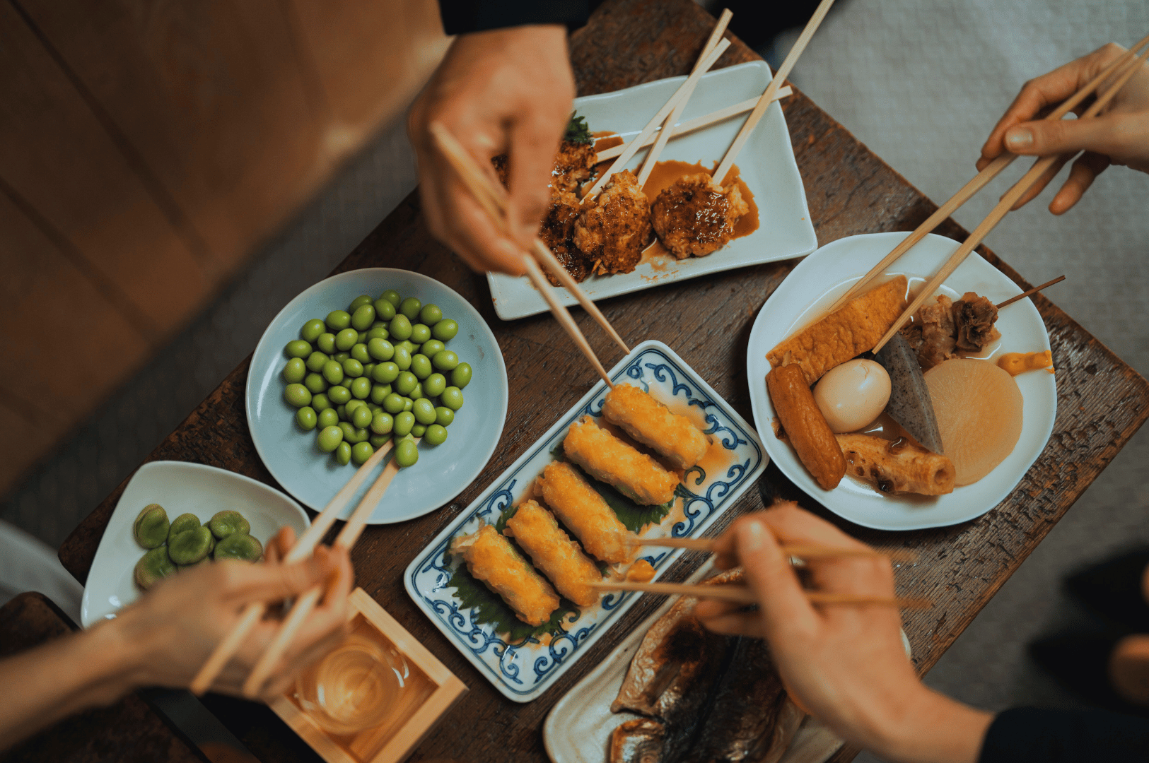 Top-down view of shared vegetarian Japanese izakaya dishes on a wooden table, with hands using chopsticks to enjoy plant-based skewers, edamame, oden, and pickles—showcasing communal dining in Singapore’s meat-free Japanese eateries.