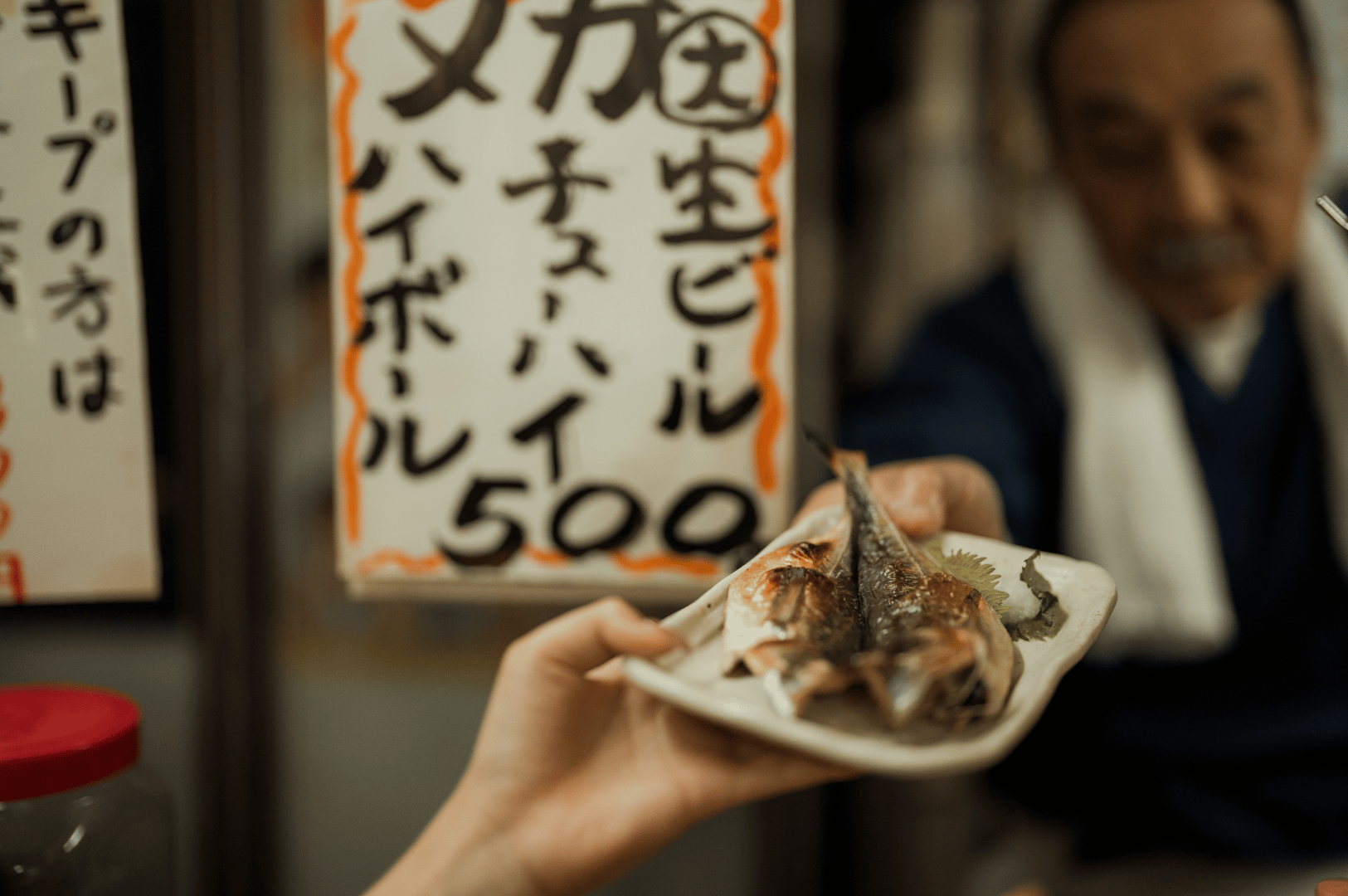 Close-up of a grilled plant-based fish dish being handed over at a Japanese food stall, with visible garnishes and a handwritten sign—capturing authentic street-style vegetarian izakaya fare in Singapore.