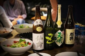 This close-up image features four bottles of Japanese sake lined up on a dark wooden table, with various bowls of salad and food items blurred in the foreground and background.