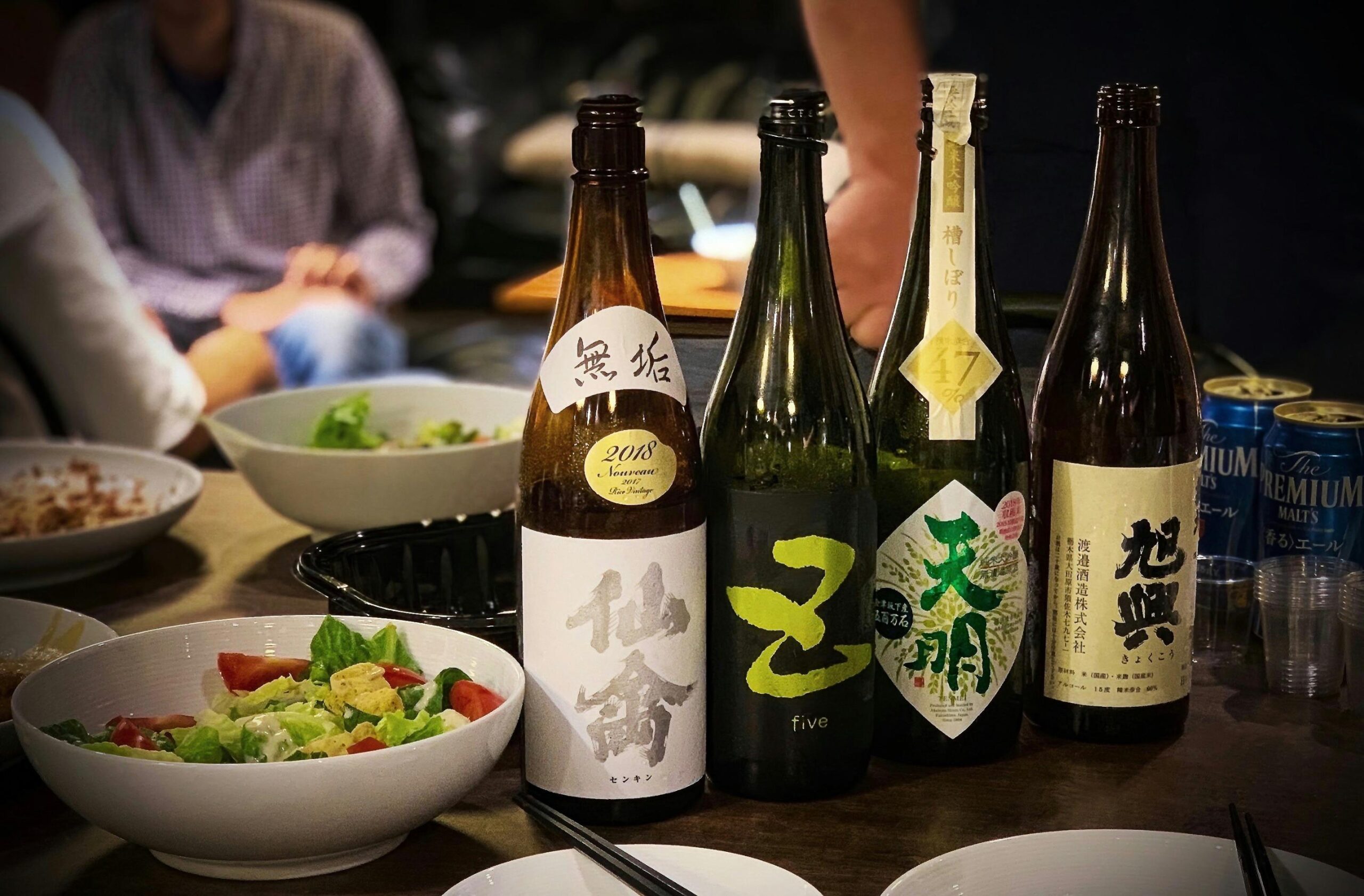 This close-up image features four bottles of Japanese sake lined up on a dark wooden table, with various bowls of salad and food items blurred in the foreground and background.