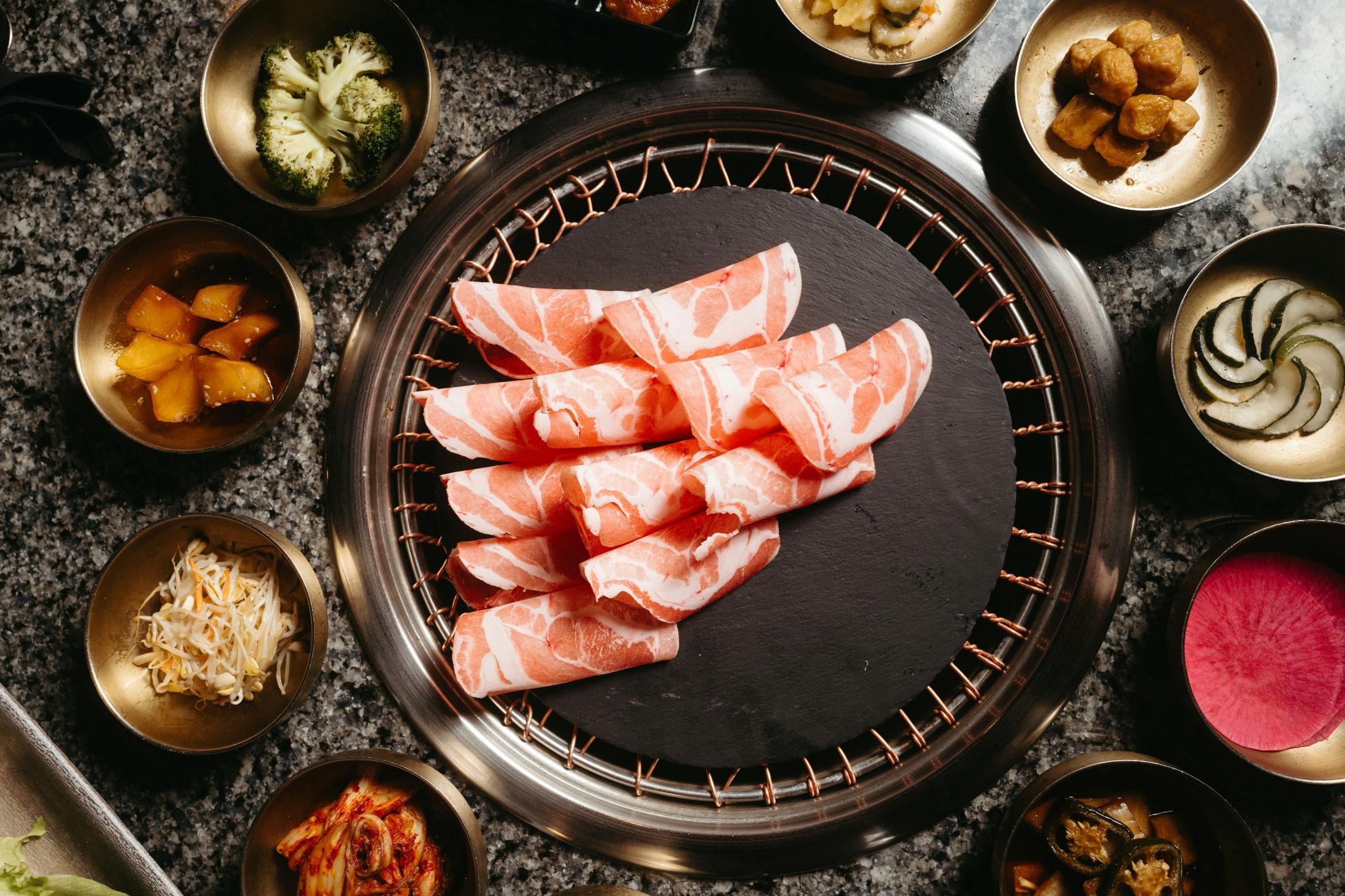 Thinly sliced rolls of raw meat are neatly arranged on a round black stone placed centrally on a wire grill. Surrounding the main dish is a colorful spread of Korean side dishes in small brass bowls, including kimchi, pickled radish, and broccoli.