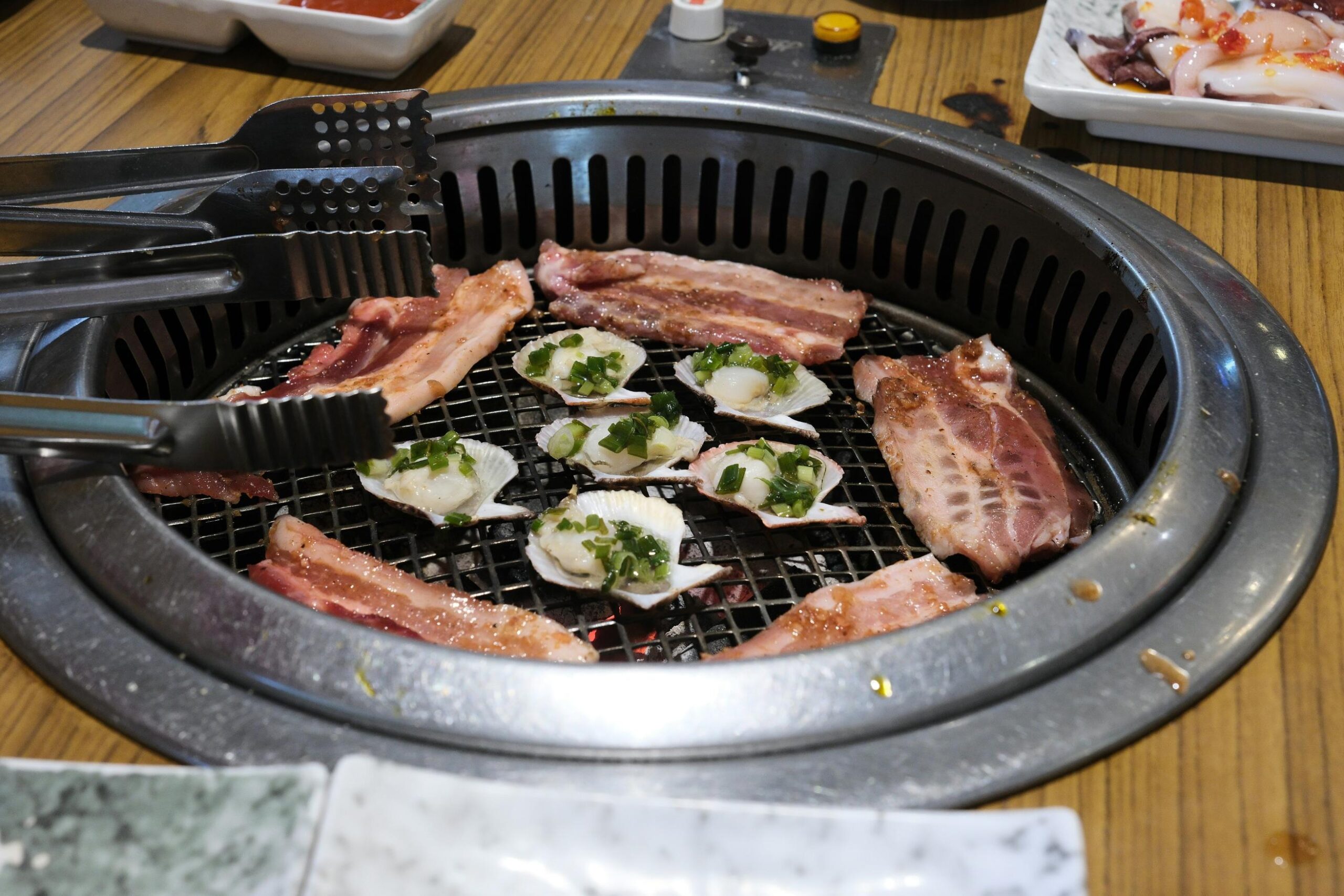 Slices of meat and scallops in their shells, garnished with chopped green onions, are cooking on a circular wire grill set into a wooden table. A pair of metal tongs reaches towards the food, capturing the interactive nature of this barbecue dining experience.