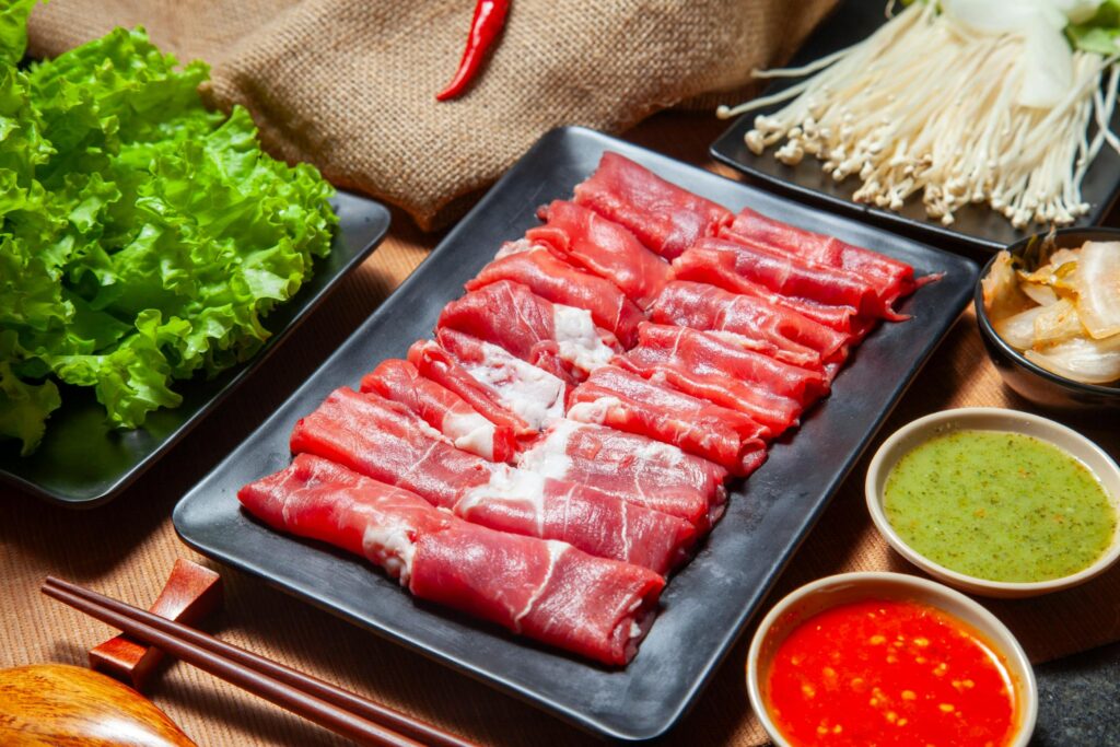 Neatly rolled slices of fresh, raw beef are arranged on a black rectangular plate, flanked by piles of green lettuce and enoki mushrooms. The scene is set for a meal with wooden utensils and small bowls of vibrant red and green dipping sauces ready for dipping.