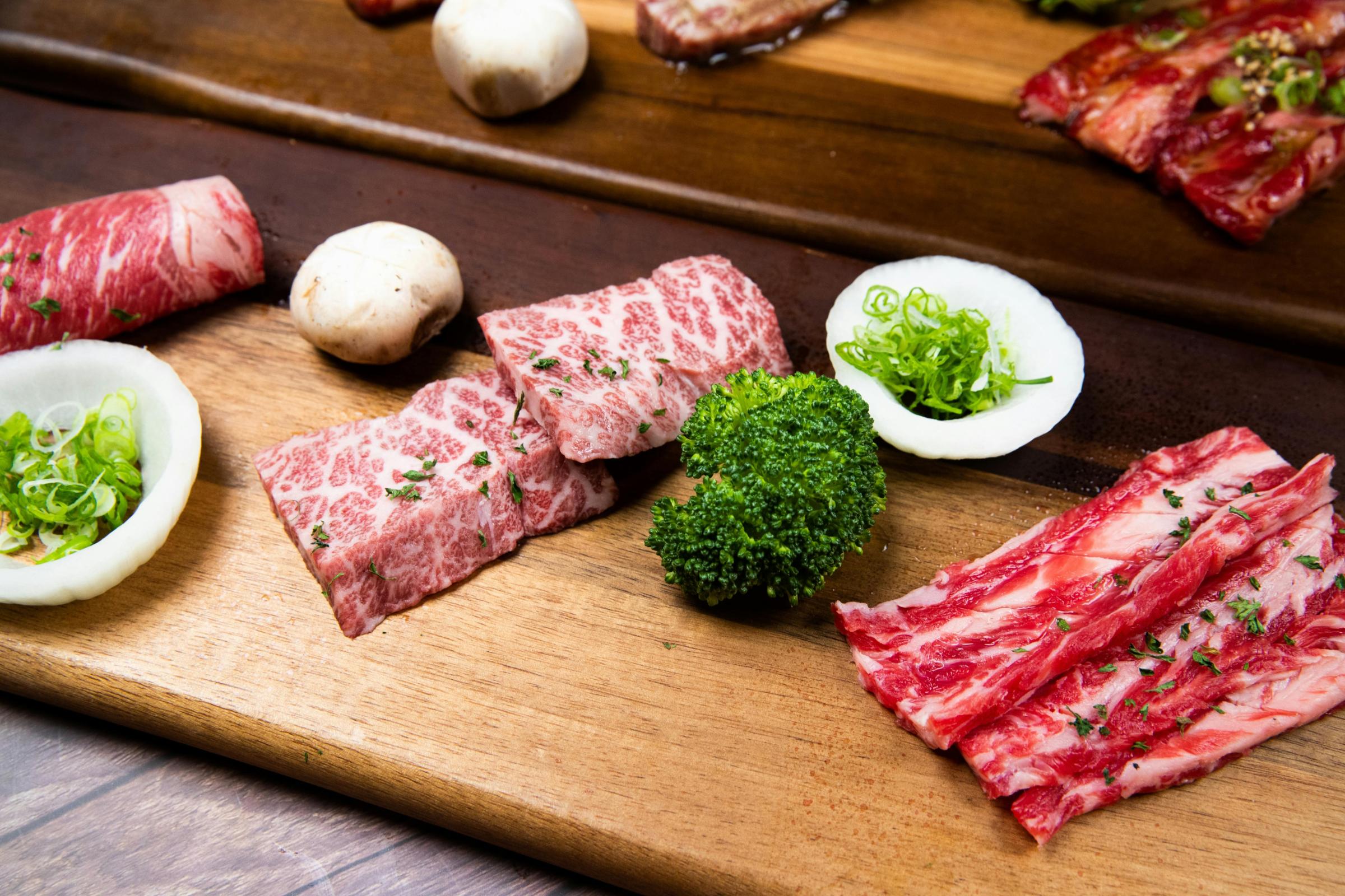 Various cuts of premium raw beef, including thick marbled cubes and tender slices, are displayed on a long wooden serving board. Fresh vegetables like whole button mushrooms, broccoli, and chopped scallions nestled in onion rings accompany the meat, ready for the grill.