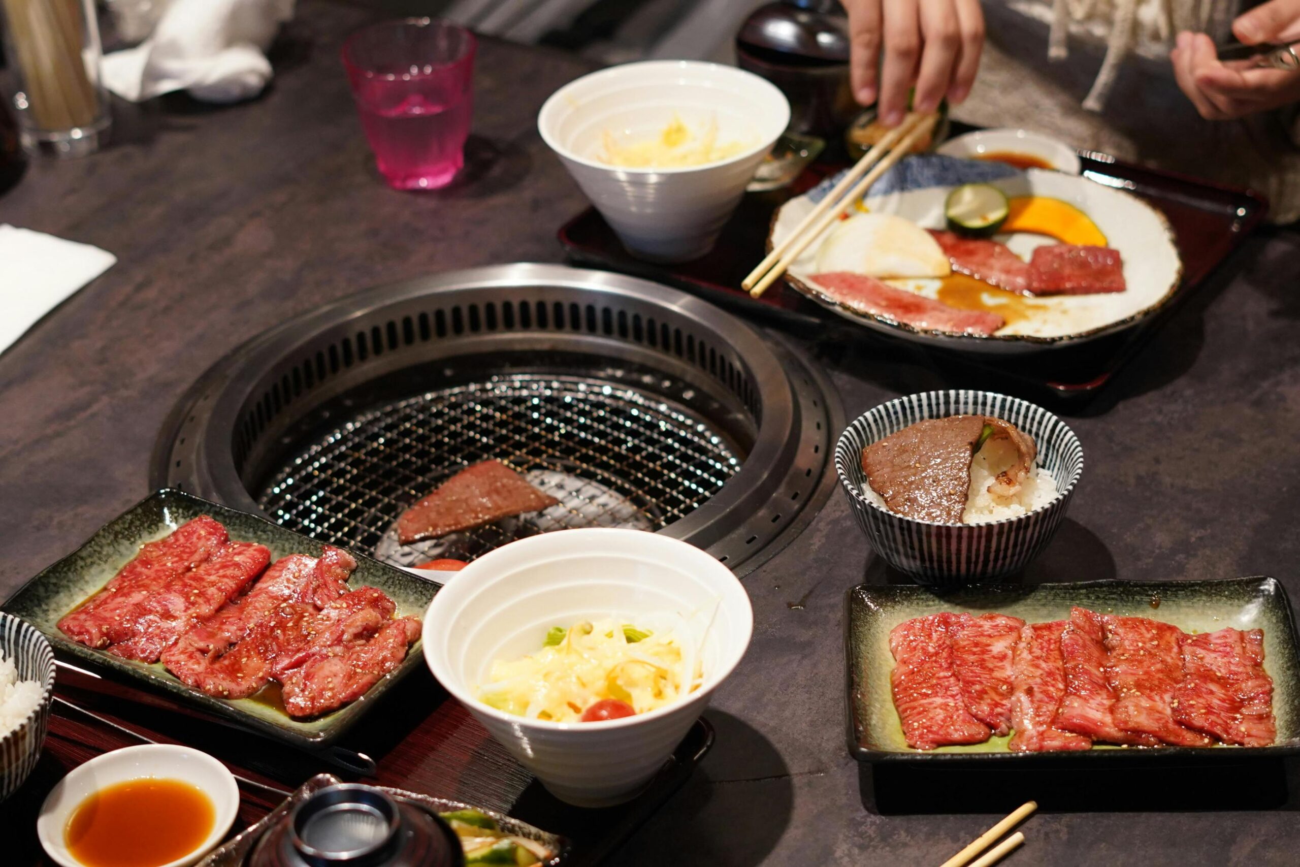 Diners enjoy a meal at a table set with plates of raw marinated meat, salads, and rice bowls topped with grilled beef. A central round wire grill cooks a slice of meat, surrounded by individual serving trays and dipping sauces.