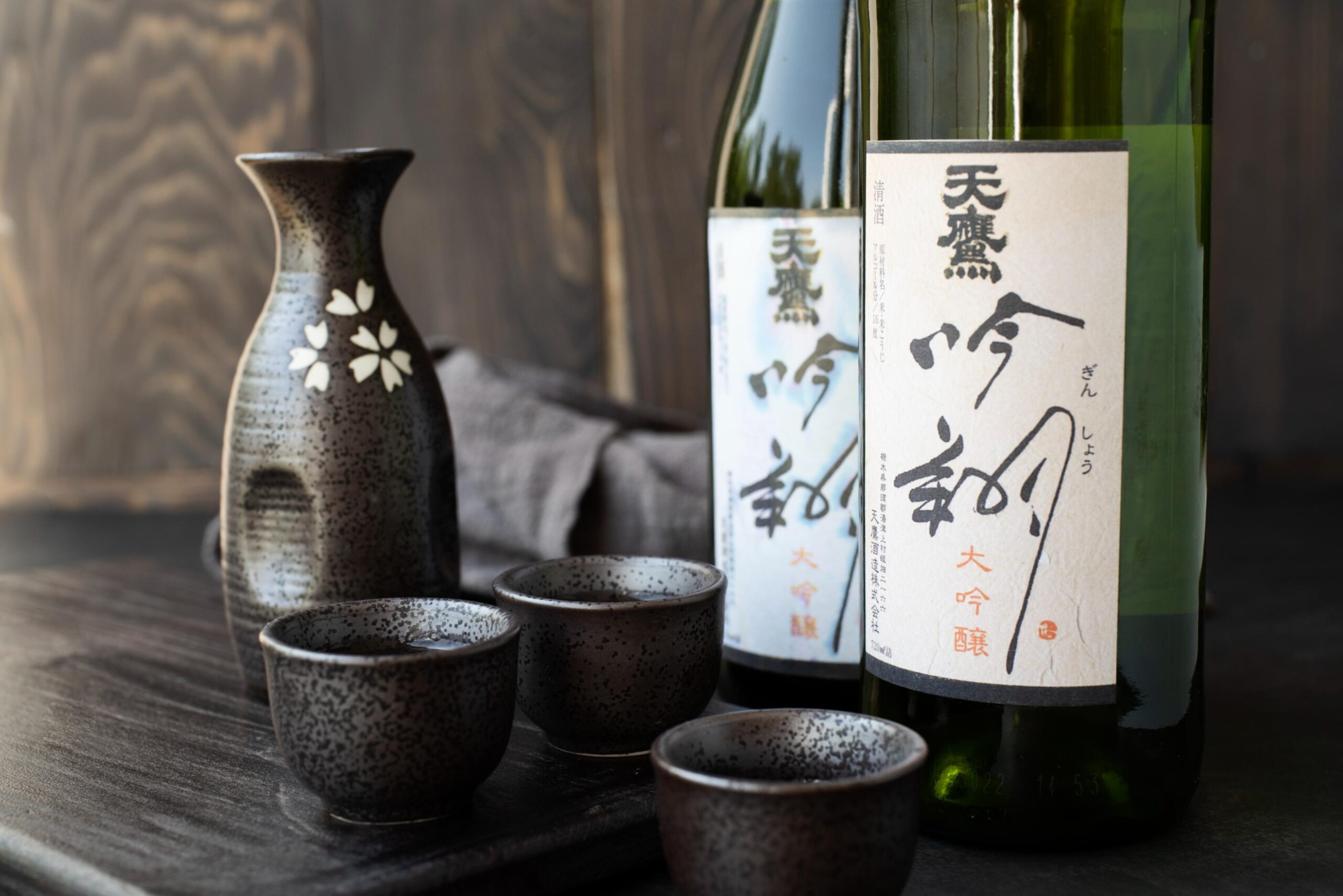 A row of ten dark glass sake bottles stands neatly on a wooden shelf against a pale wall accented with vertical green stripes. While several bottles feature matching black labels with white calligraphy, a single bottle near the right stands out with a bright white label.