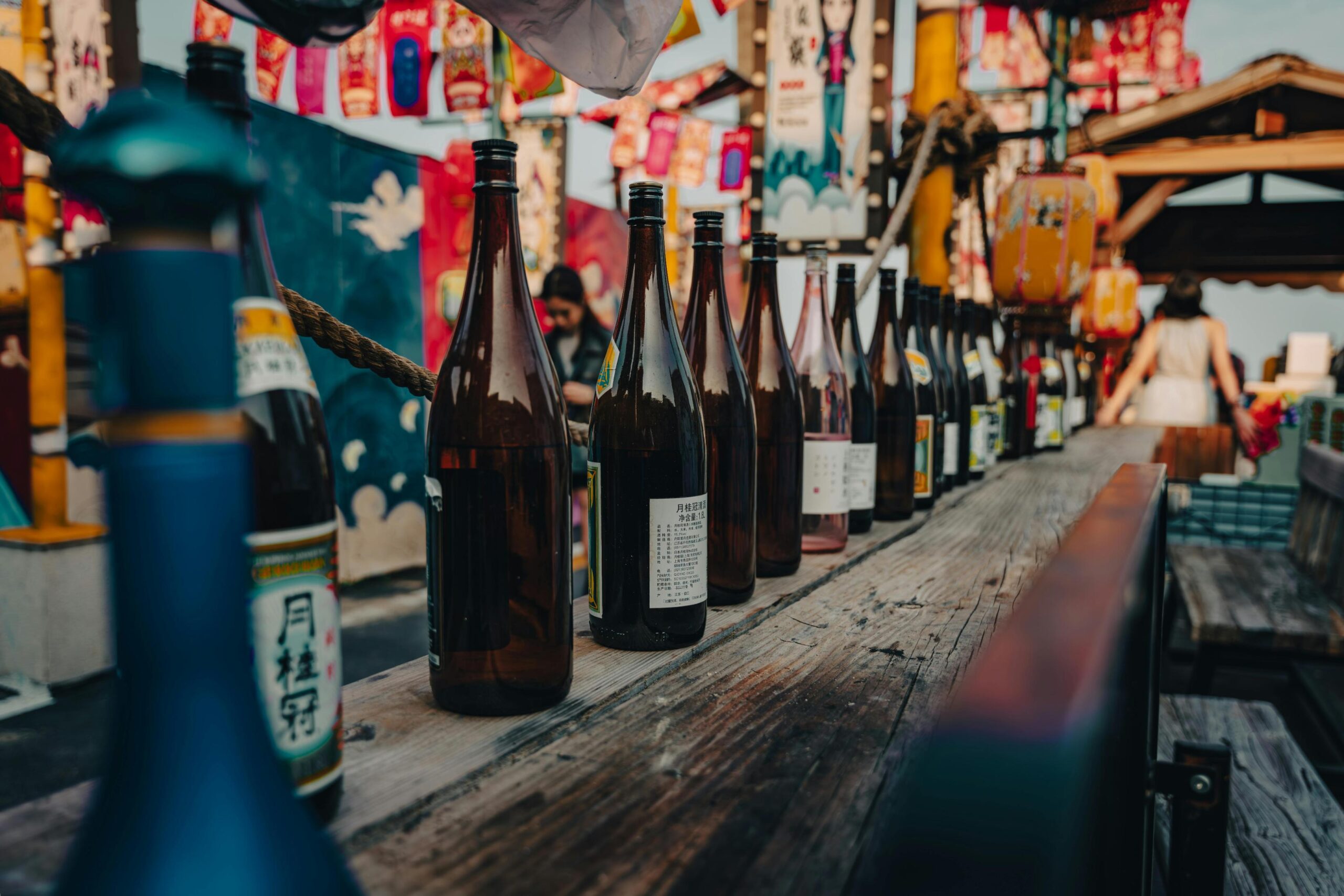 A textured ceramic tokkuri with white floral accents sits alongside three matching ochoko cups on a dark, rustic surface. Behind them, two green glass sake bottles featuring elegant cream labels are displayed against a warm, wood-grained background.