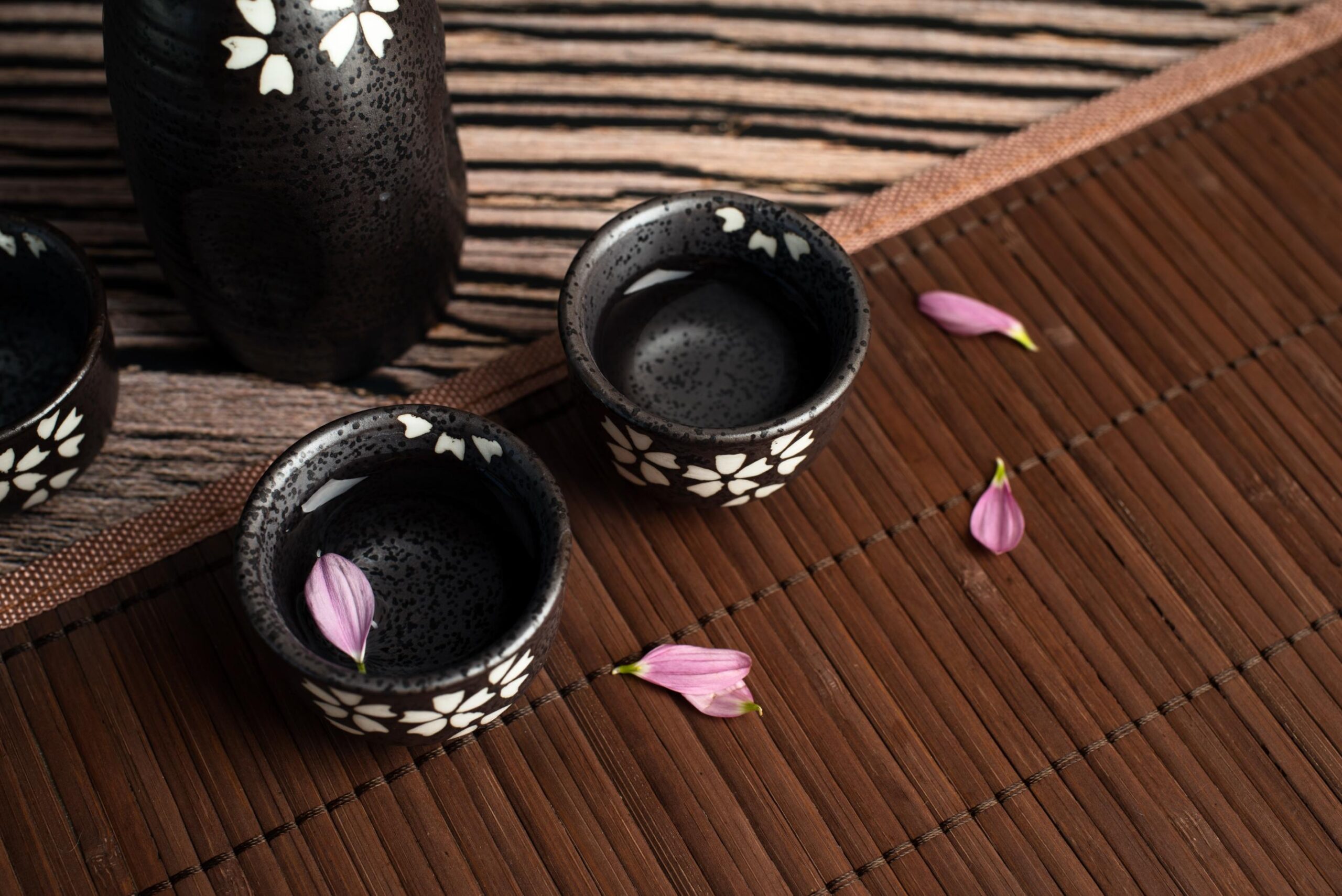 This high-angle shot features two dark ceramic ochoko cups and a matching bottle, all decorated with delicate white flower patterns. Pink flower petals are scattered across a brown bamboo mat and inside one of the cups, adding a soft touch to the rustic wooden setting.