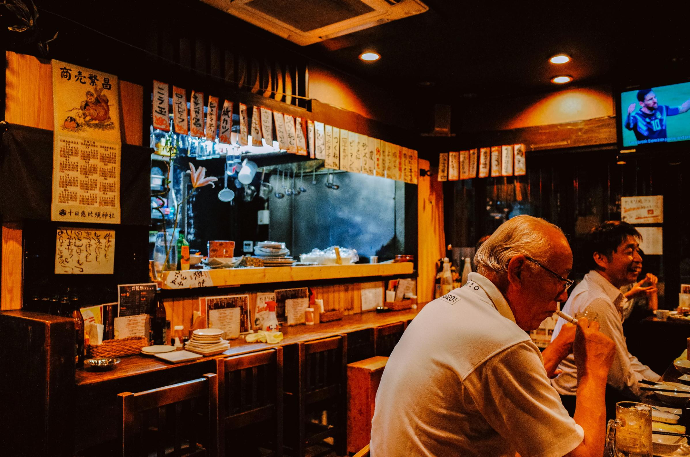 This candid shot captures the warm atmosphere of a traditional Japanese izakaya, featuring an older man dining at a wooden counter while a soccer match plays on a television in the corner. Rows of handwritten paper menus hang above the open kitchen area, adding authentic detail to the cozy, dimly lit interior.