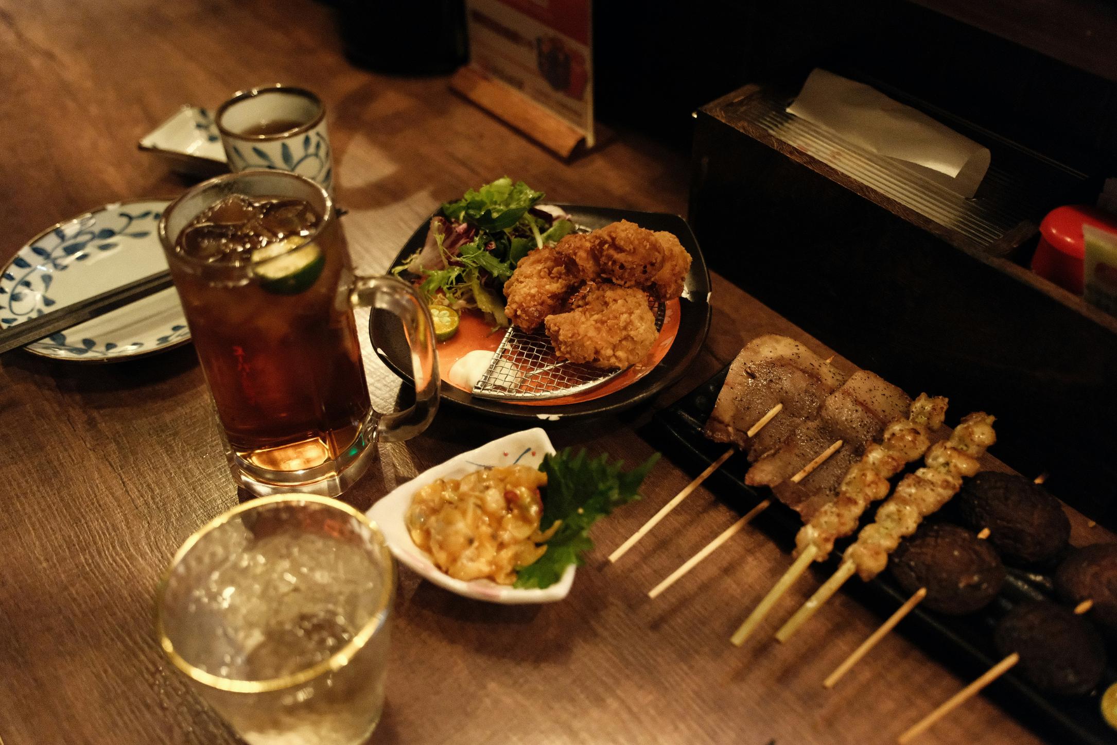 A wooden table is set with a variety of Japanese dishes, including skewered grilled meats, crispy fried chicken, small side plates, and fresh greens. Drinks like iced tea and a clear spirit sit alongside the food, creating a cozy, inviting izakaya-style dining scene.