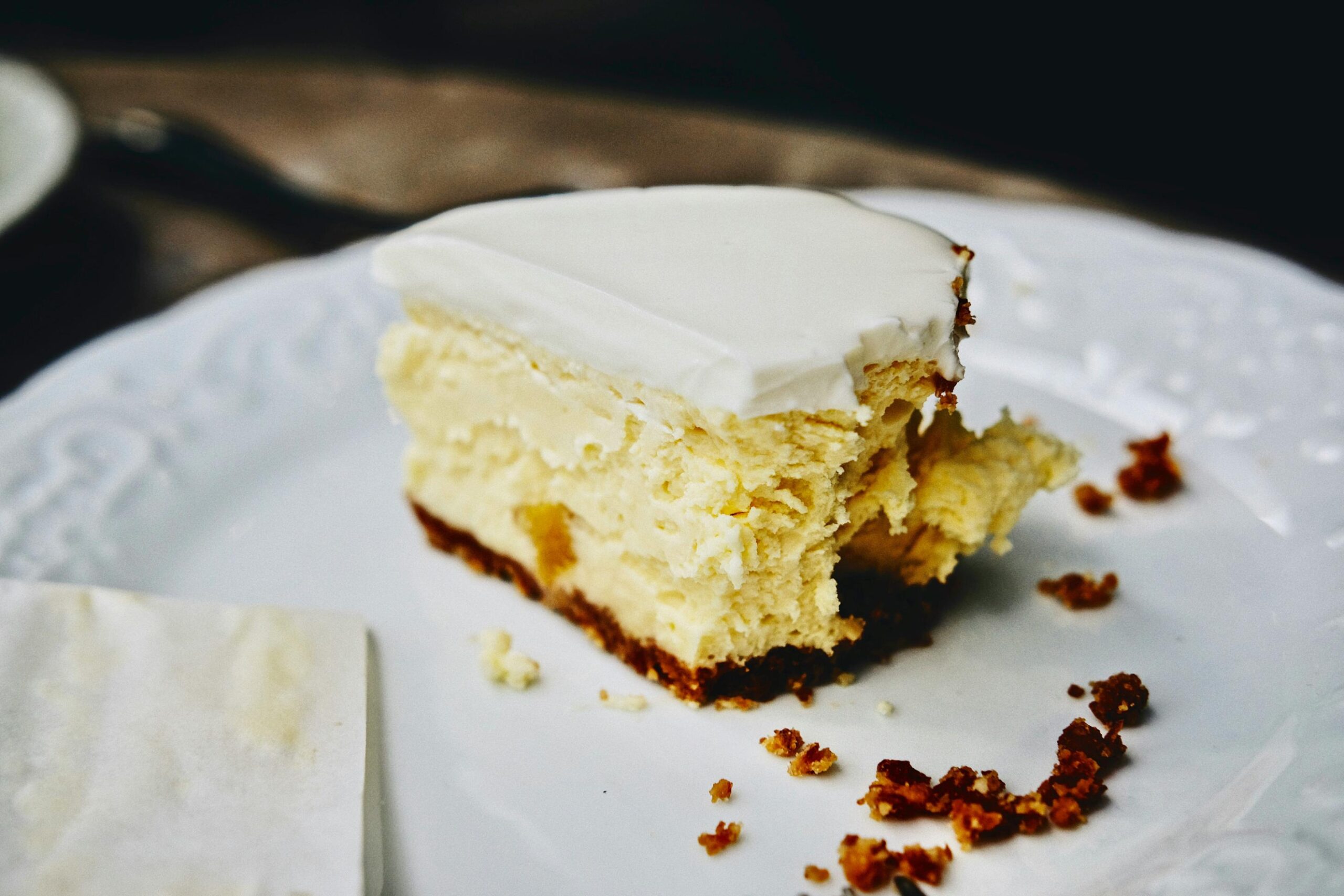A generous slice of cheesecake with a smooth white topping rests on an embossed plate, featuring a dark crumb crust. The dessert appears partially eaten, with a jagged edge and scattered crumbs decorating the white surface.