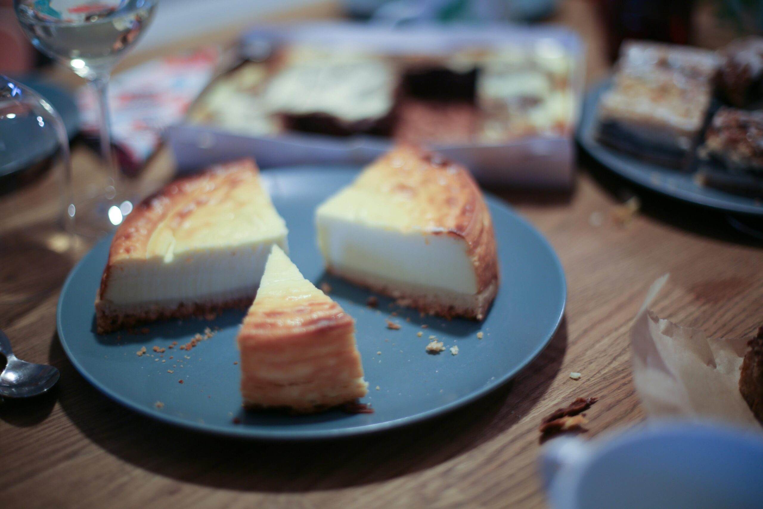 Three slices of golden-brown cheesecake rest on a blue plate in the foreground of a wooden table. The background remains out of focus, revealing a casual dessert spread with a wine glass and additional trays of pastries.