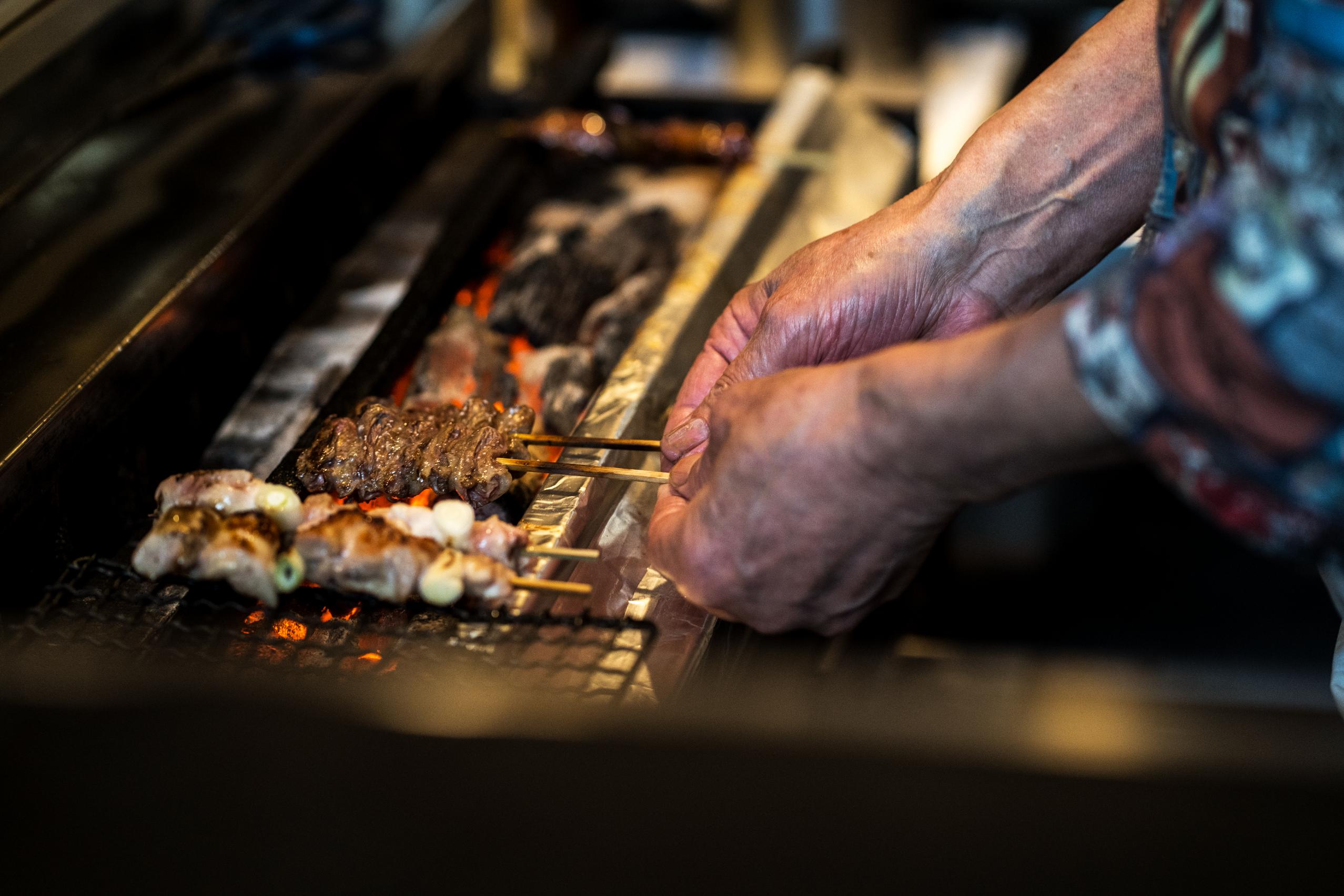 A cook's hands carefully adjust skewers of meat grilling over hot coals, with the glowing embers visible beneath the metal grate. The high-angle close-up captures the smoky atmosphere and the texture of the street food as it cooks.