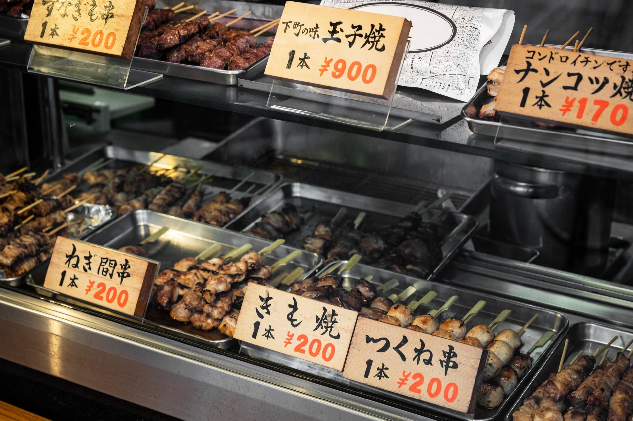 Metal trays filled with various grilled skewers are arranged inside a glass display case, showcasing a selection of Japanese street food. Wooden signs featuring Japanese text and prices in Yen sit in front of the trays to identify the different options available for purchase.
