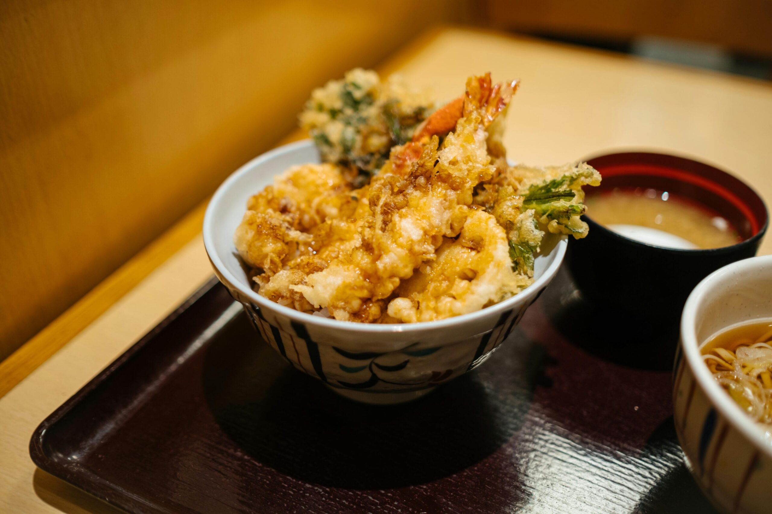 This image features a hearty bowl of tendon, consisting of assorted crispy tempura such as shrimp and vegetables drizzled with a savory sauce over rice. The meal is presented on a traditional dark tray alongside a small bowl of miso soup and a side of noodles, offering a classic Japanese comfort food experience.