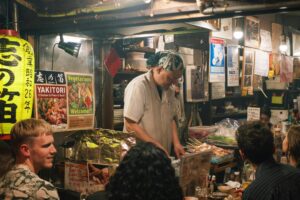 In a cluttered and cozy Japanese eatery, a focused chef wearing a headband prepares food behind a counter lined with posters and a "Yakitori" sign. Customers sit at the crowded bar in the foreground, illuminated by the warm glow of a large yellow paper lantern and overhead lights.