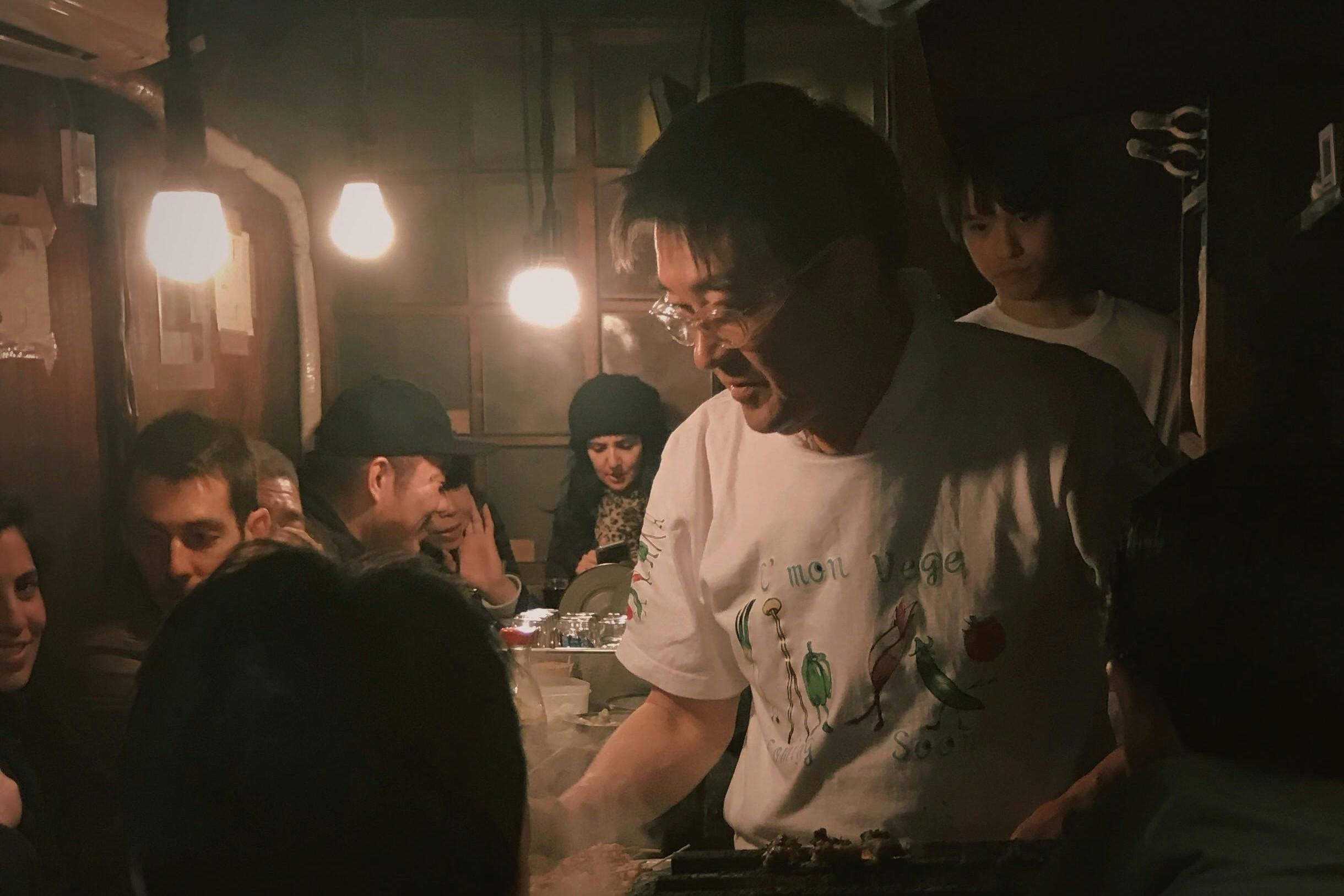 In a dimly lit and smoky restaurant, a chef wearing glasses and a white t-shirt grills food beneath the warm glow of hanging light bulbs. Patrons are seated closely around the cooking area, observing the chef in the intimate and atmospheric setting.