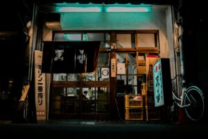 This nighttime shot depicts the rustic storefront of a Japanese restaurant, characterized by traditional noren curtains hanging over sliding wooden doors and vertical signage. Stacks of yellow Kirin beer crates and a parked bicycle line the entrance, bathed in the cool glow of the overhead fluorescent lights.