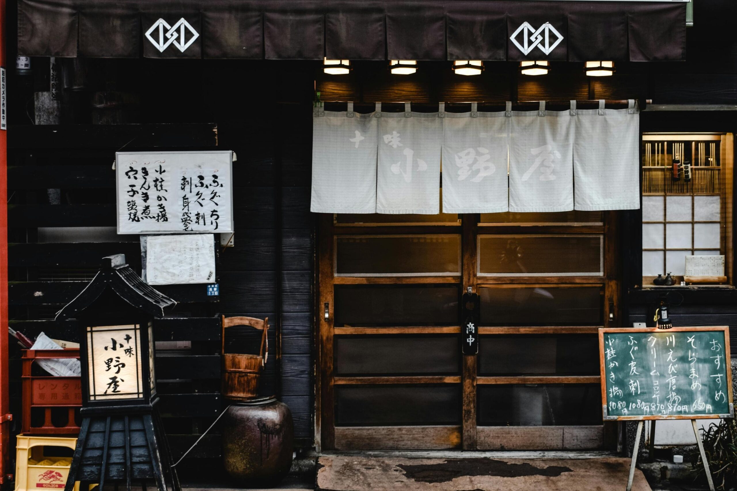 This image captures the traditional dark wooden facade of a Japanese eatery, featuring sliding doors draped with white fabric noren curtains. The entrance is flanked by an illuminated paper lantern stand on the left and a handwritten chalkboard menu on the right, creating a rustic and inviting atmosphere.