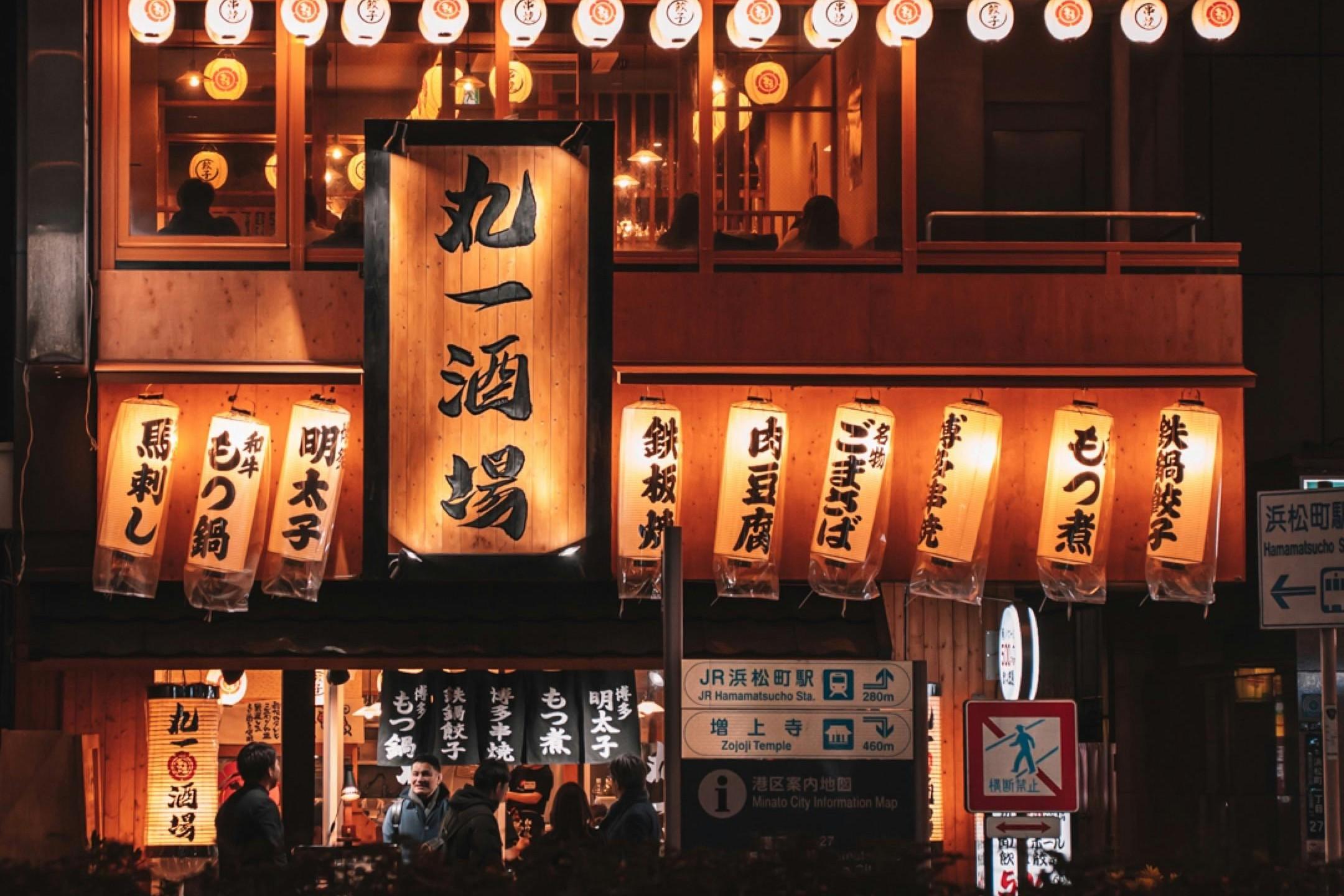 This night scene showcases the vibrant exterior of a multi-story Japanese restaurant, distinguished by a massive vertical wooden sign and rows of glowing paper lanterns hanging across the facade. Below the warm lights, people gather near the entrance, while street signs indicate the location is near JR Hamamatsucho Station and Zojoji Temple.