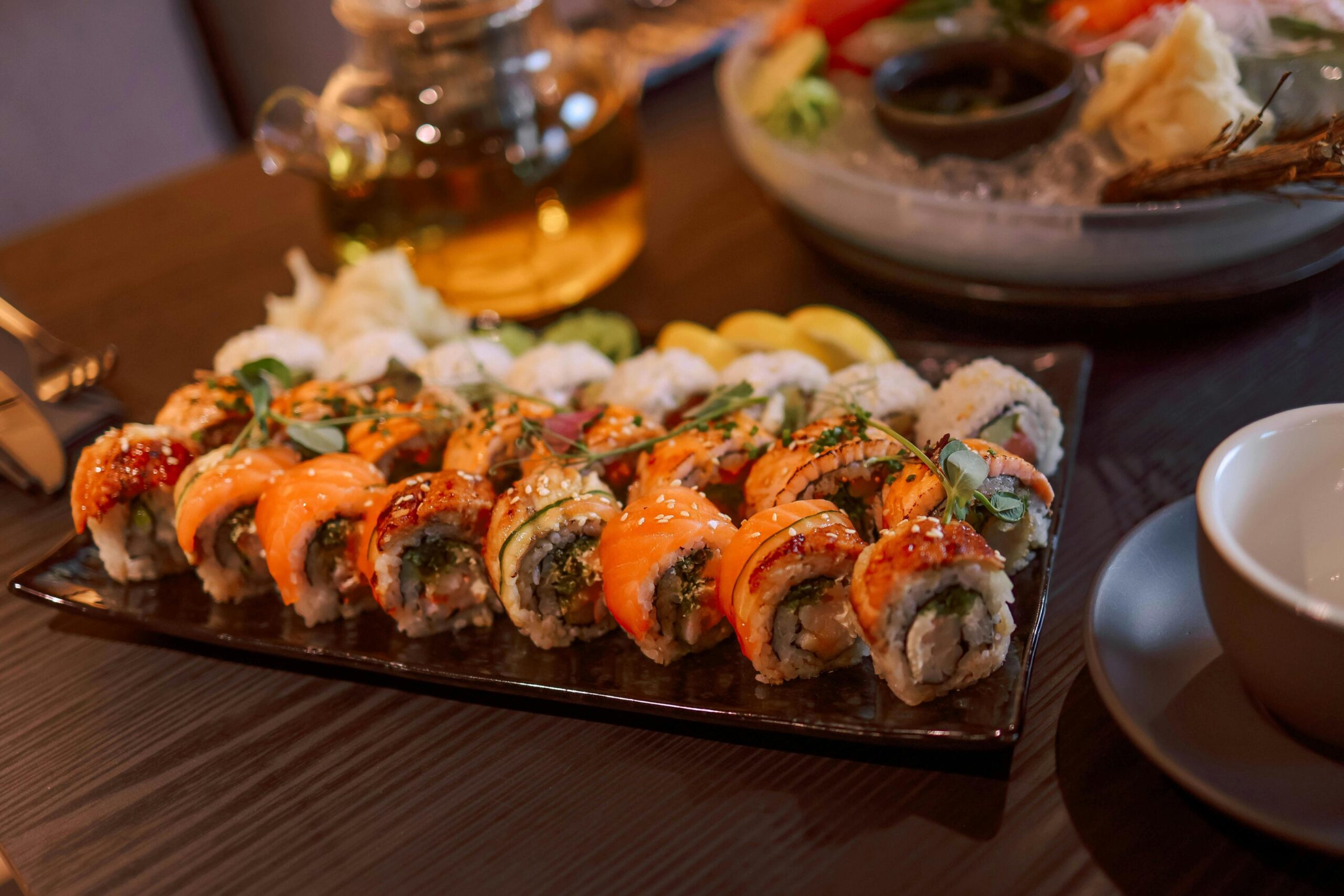 A dark rectangular platter presents a delicious array of sushi rolls topped with fresh salmon and glazed fish, garnished with sesame seeds and microgreens. The warm, inviting scene also features a glass teapot filled with amber tea and a glimpse of a sashimi dish in the background.