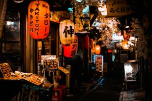 Glowing red paper lanterns emblazoned with "YAKITORI" and Japanese characters hang densely overhead, casting a warm light over a narrow, shadowy alleyway. The scene is decorated with artificial white foliage and lined with menu stands, creating the moody and inviting atmosphere of a traditional Japanese dining street.
