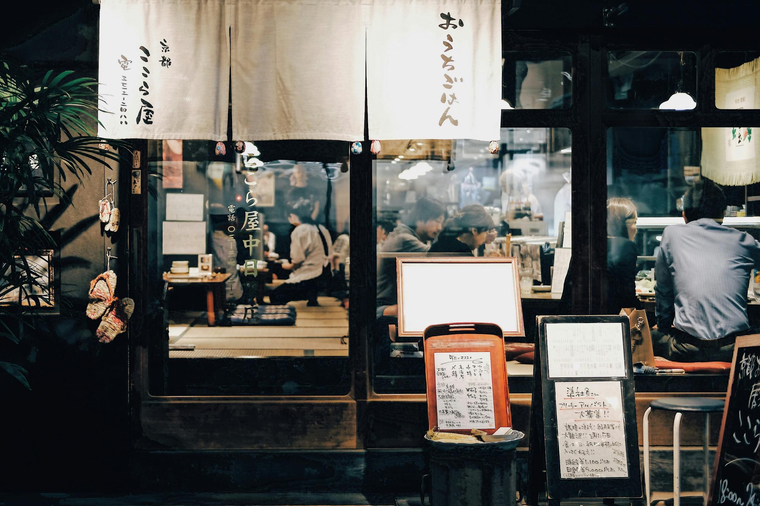 This image captures the inviting storefront of a traditional Japanese restaurant at night, featuring white noren curtains with calligraphy and handwritten menu boards standing near the entrance. Through the large glass windows, a warm and lively interior is visible, showing patrons dining at counters and low tables in a cozy atmosphere.
