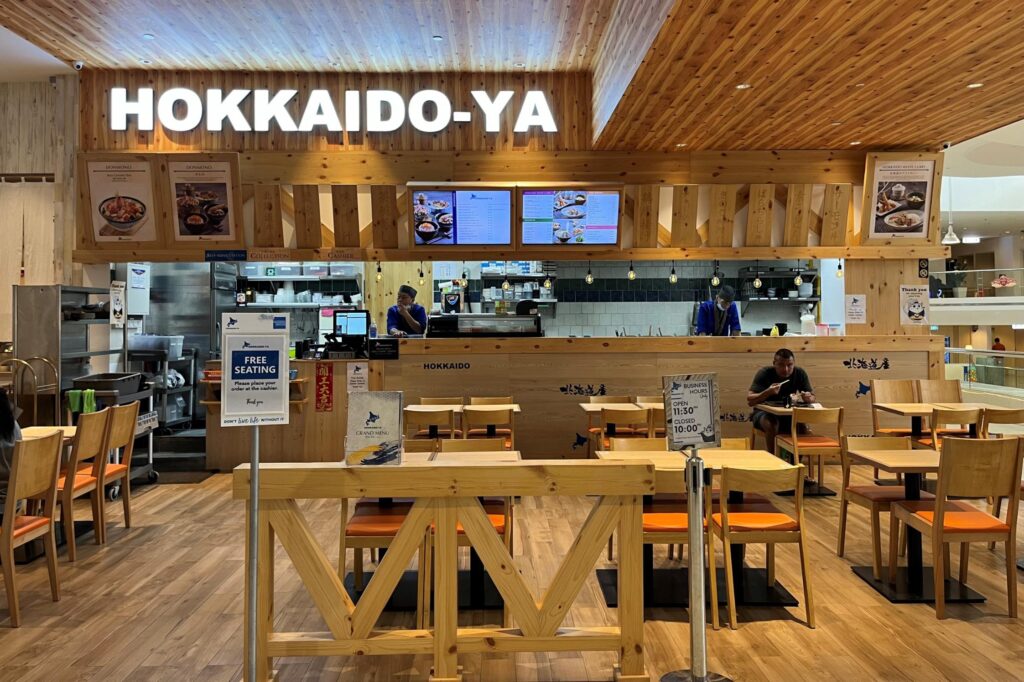 The image shows the brightly lit storefront of Hokkaido-Ya, a Japanese restaurant featuring light wood paneling and a clean, modern aesthetic. Several wooden tables and chairs are arranged in the foreground for diners, with staff visible behind the service counter and digital menu boards displayed above.