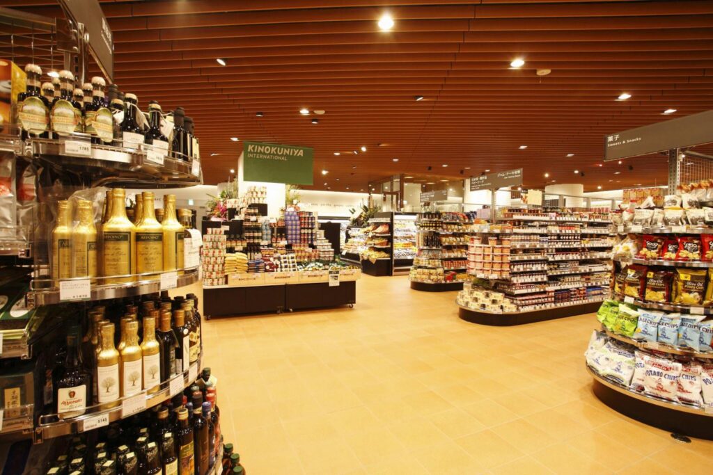 The image showcases the spacious interior of a Kinokuniya International grocery store, featuring polished floors and a distinctive slatted wooden ceiling. Neatly organized curved shelves and island displays are stocked with various food products, including snacks, oils, and bottled beverages.