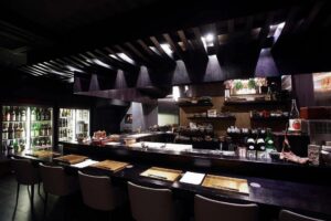 An elegant, dimly lit Japanese sake bar features a long dark countertop set with gold-colored placemats and neutral-toned chairs. Behind the bar, shelves of artisanal pottery and a large refrigerated display filled with sake bottles create a sophisticated, moody atmosphere.