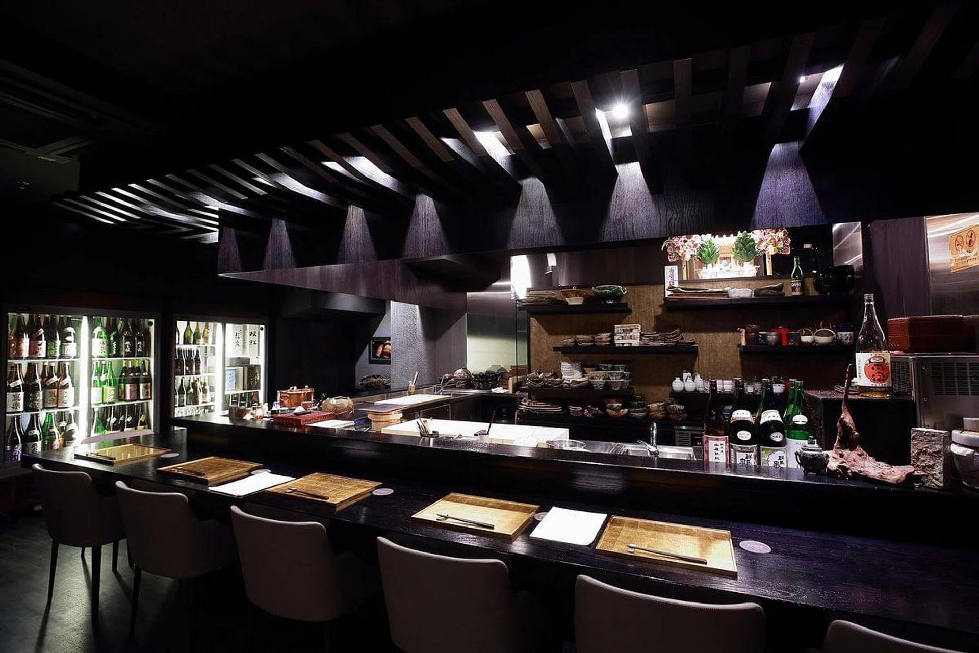 An elegant, dimly lit Japanese sake bar features a long dark countertop set with gold-colored placemats and neutral-toned chairs. Behind the bar, shelves of artisanal pottery and a large refrigerated display filled with sake bottles create a sophisticated, moody atmosphere.