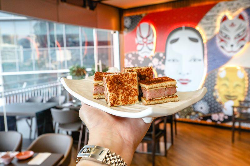 A person holds a white plate of toasted sandwiches featuring thick, pink fillings in a modern restaurant setting. The background showcases large, colorful Japanese-inspired wall art and a bright window view of an urban area.