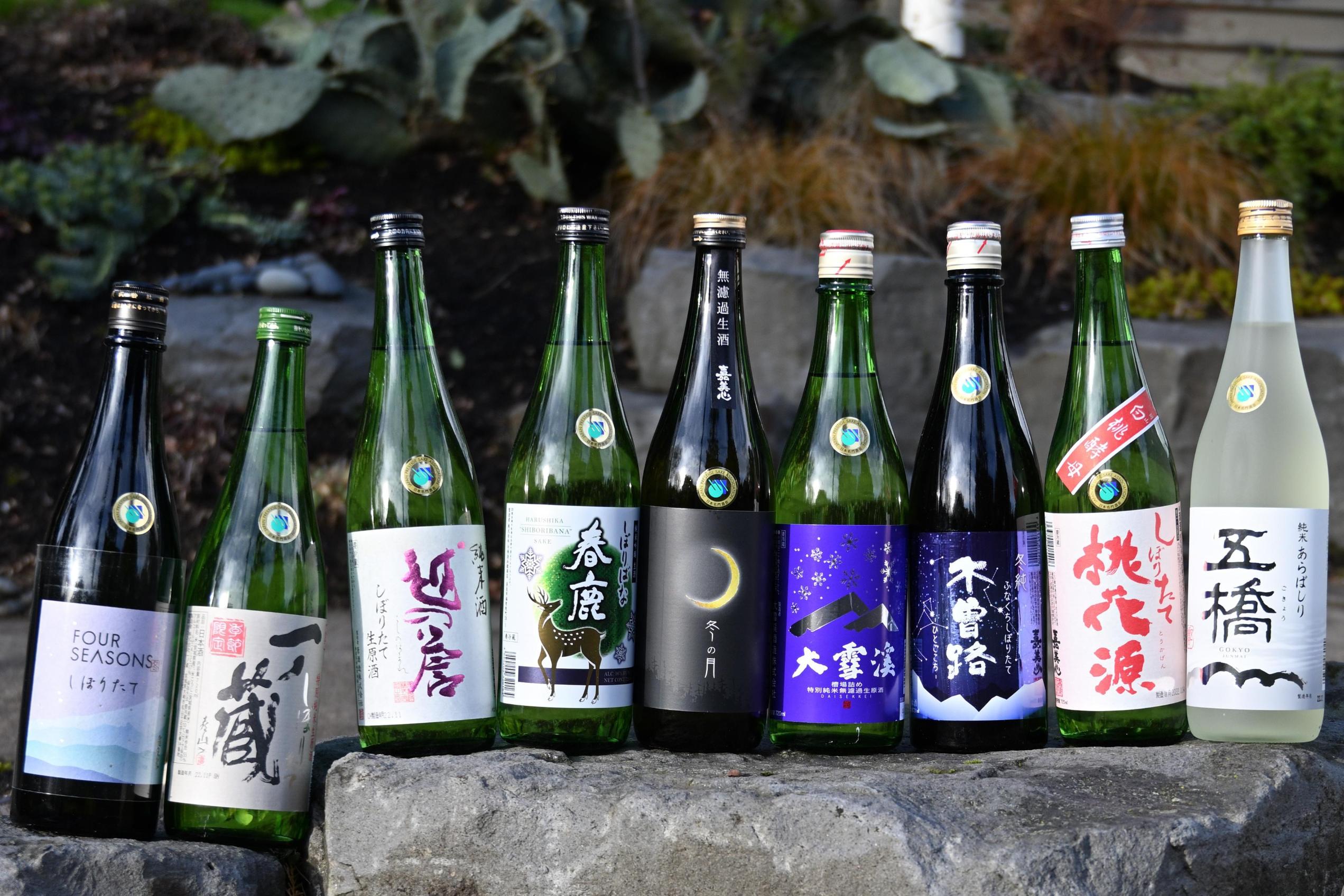 Nine bottles of Japanese sake, featuring diverse colorful labels and various bottle colors, are lined up in a row on a grey stone ledge. The background shows a blurred outdoor garden setting with green plants and mulch, highlighting the selection of beverages.