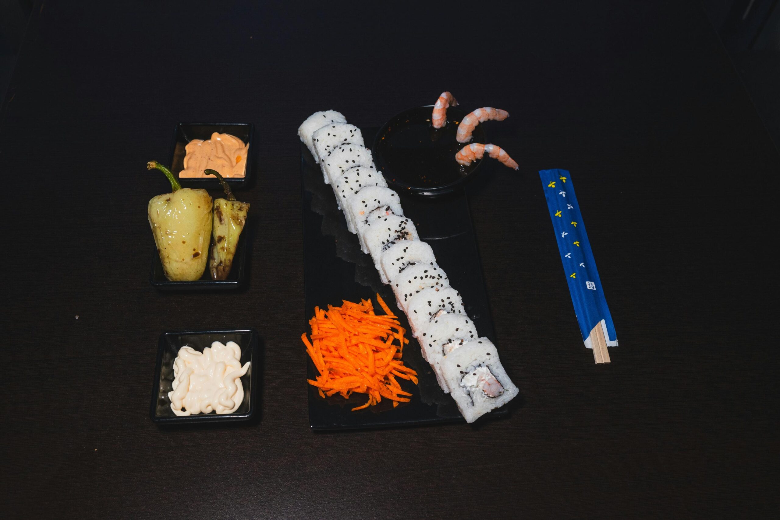 This image displays a long row of sushi rolls topped with black sesame seeds, arranged on a black platter alongside shredded carrots and a dipping bowl with shrimp. The spread is accompanied by small side dishes containing grilled peppers and various creamy sauces, all set against a dark, minimalist background.