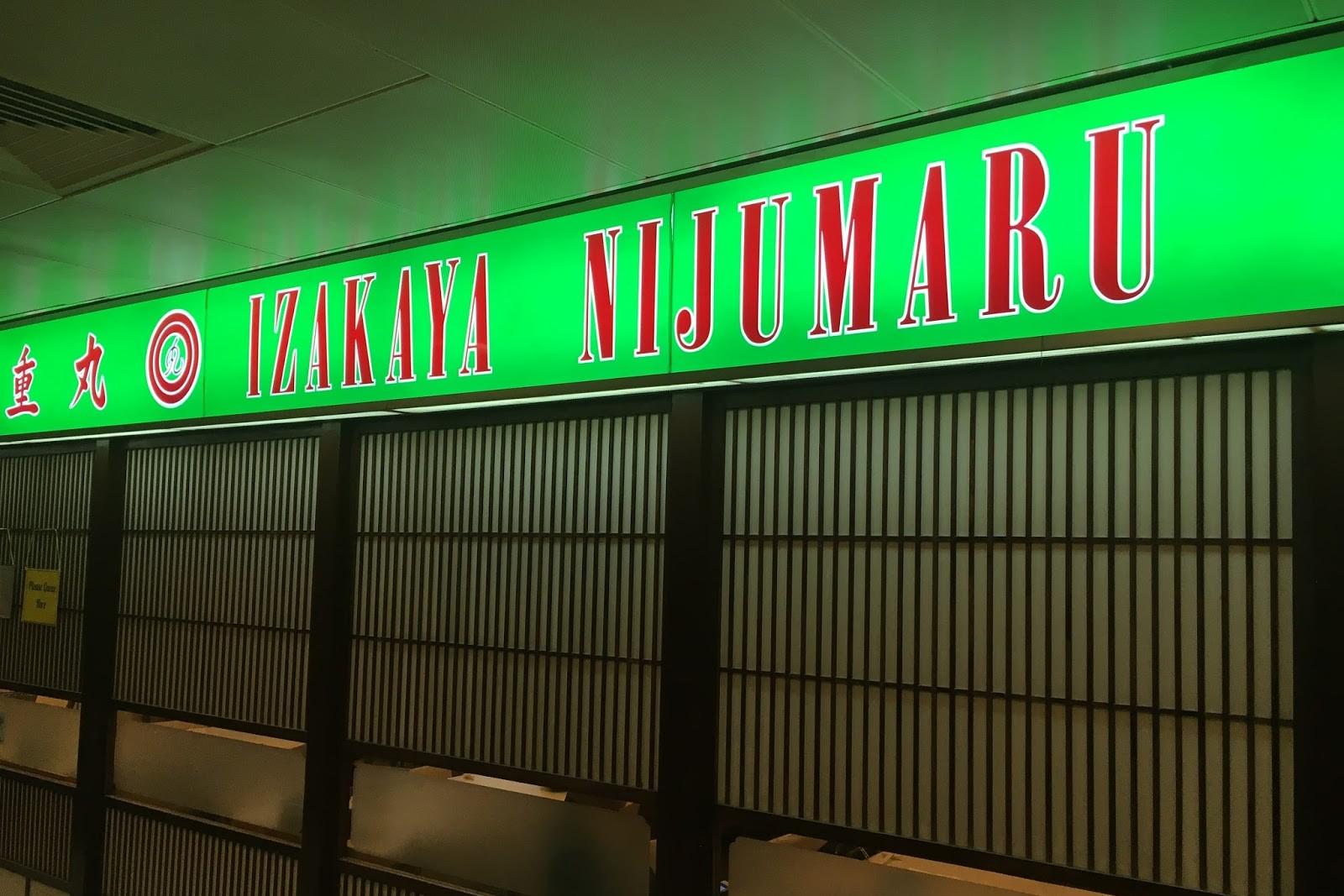 This image features the vibrant green, illuminated storefront sign for Izakaya Nijumaru, which includes the restaurant's name in both English and Japanese characters. Below the sign, traditional dark-framed sliding doors with vertical wooden slats create a classic Japanese aesthetic for the entrance.