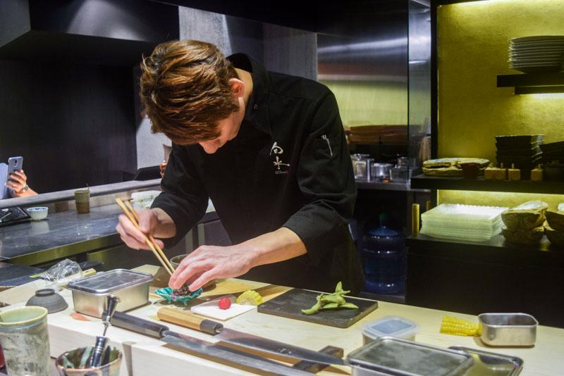 A chef in a black uniform meticulously plates small appetizers using chopsticks at a clean, well-organized wooden station. The background features a modern kitchen setting with golden-hued shelving stacked with ceramic dishes and professional culinary equipment.