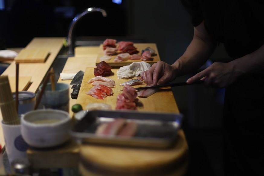 A chef’s hands are shown in close-up as they use a long, thin knife to precisely slice various cuts of fresh sashimi on a wooden block. The workspace is organized with multiple varieties of raw fish, including tuna and whitefish, awaiting preparation for service.