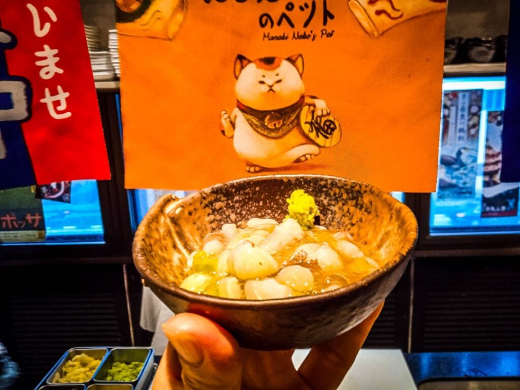A small, textured ceramic bowl filled with translucent seafood and topped with a dab of green wasabi is held up in the foreground. The background features a vibrant orange banner with an