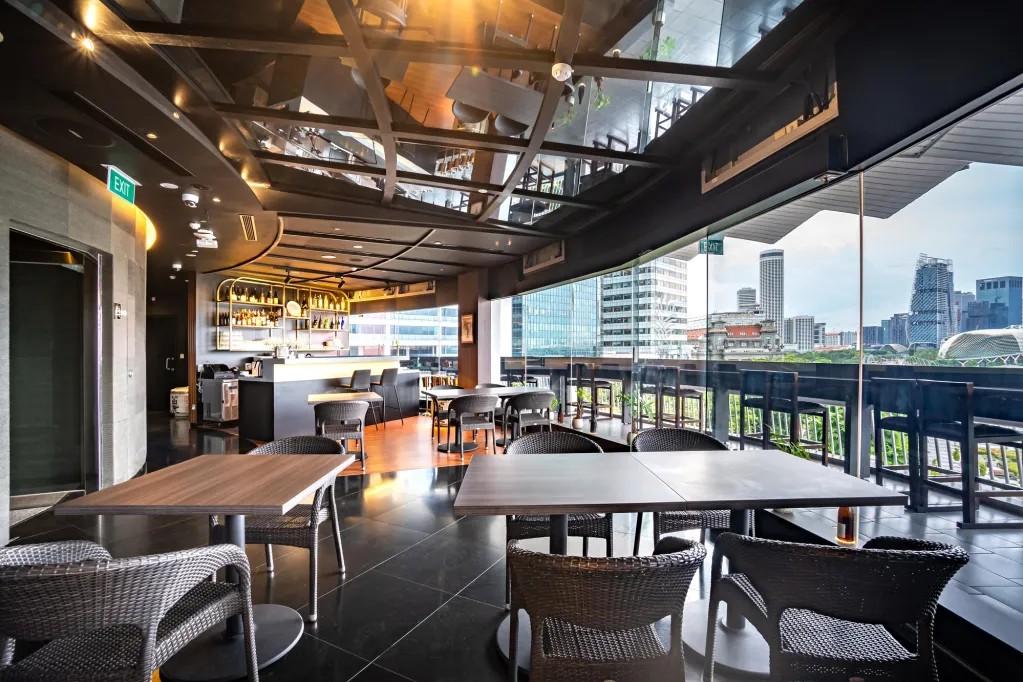 The modern interior of this restaurant features sleek black tiling and dark woven chairs arranged around simple wooden tables. Expansive floor-to-ceiling windows offer a panoramic view of a city skyline, while a well-stocked bar and reflective ceiling grid add to the sophisticated atmosphere.