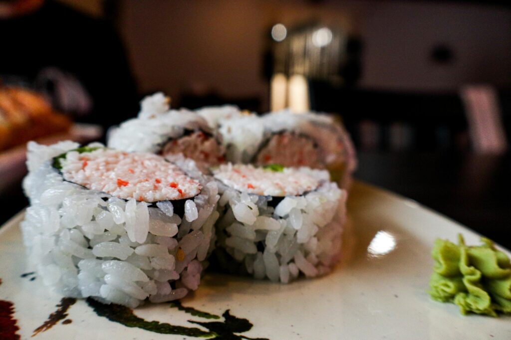 This close-up shot features several pieces of sushi rolls filled with crab salad and wrapped in white rice. A small swirl of green wasabi sits on the edge of the speckled ceramic plate in the foreground.