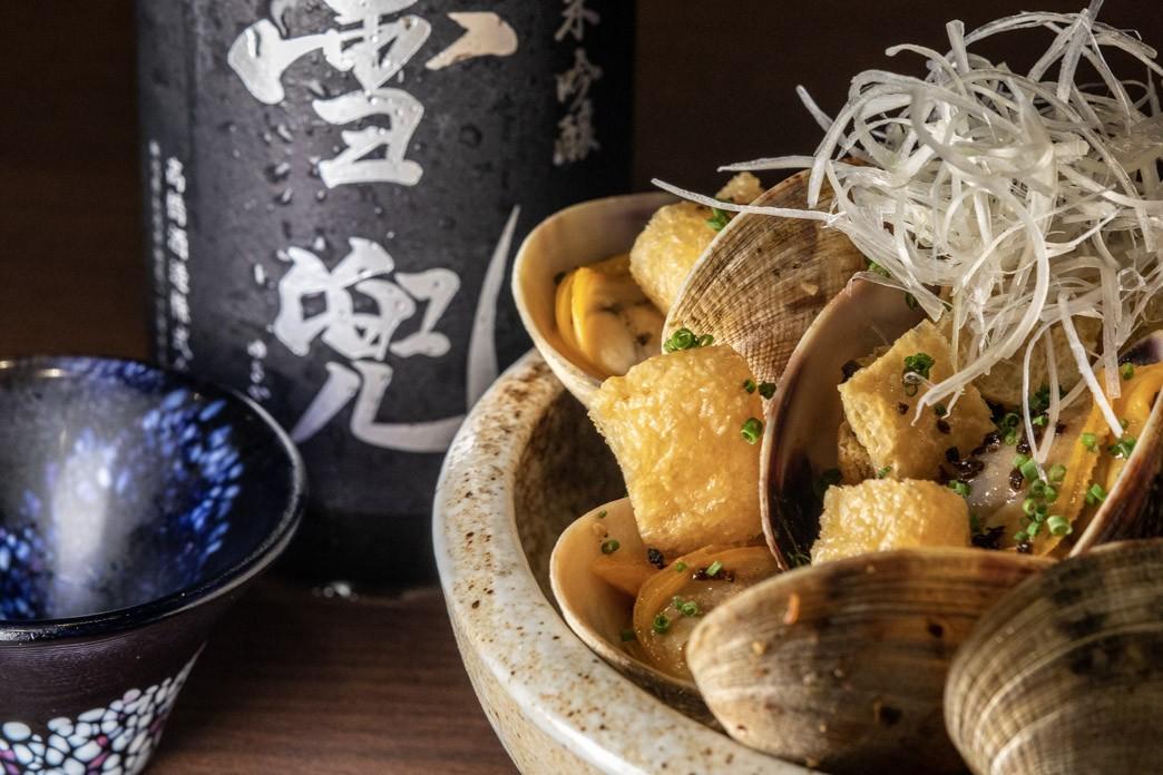 A ceramic bowl is filled with cooked clams, crispy tofu cubes, and a delicate garnish of shredded white radish. In the background, a frosted bottle of sake and a small patterned cup suggest a traditional Japanese dining experience.