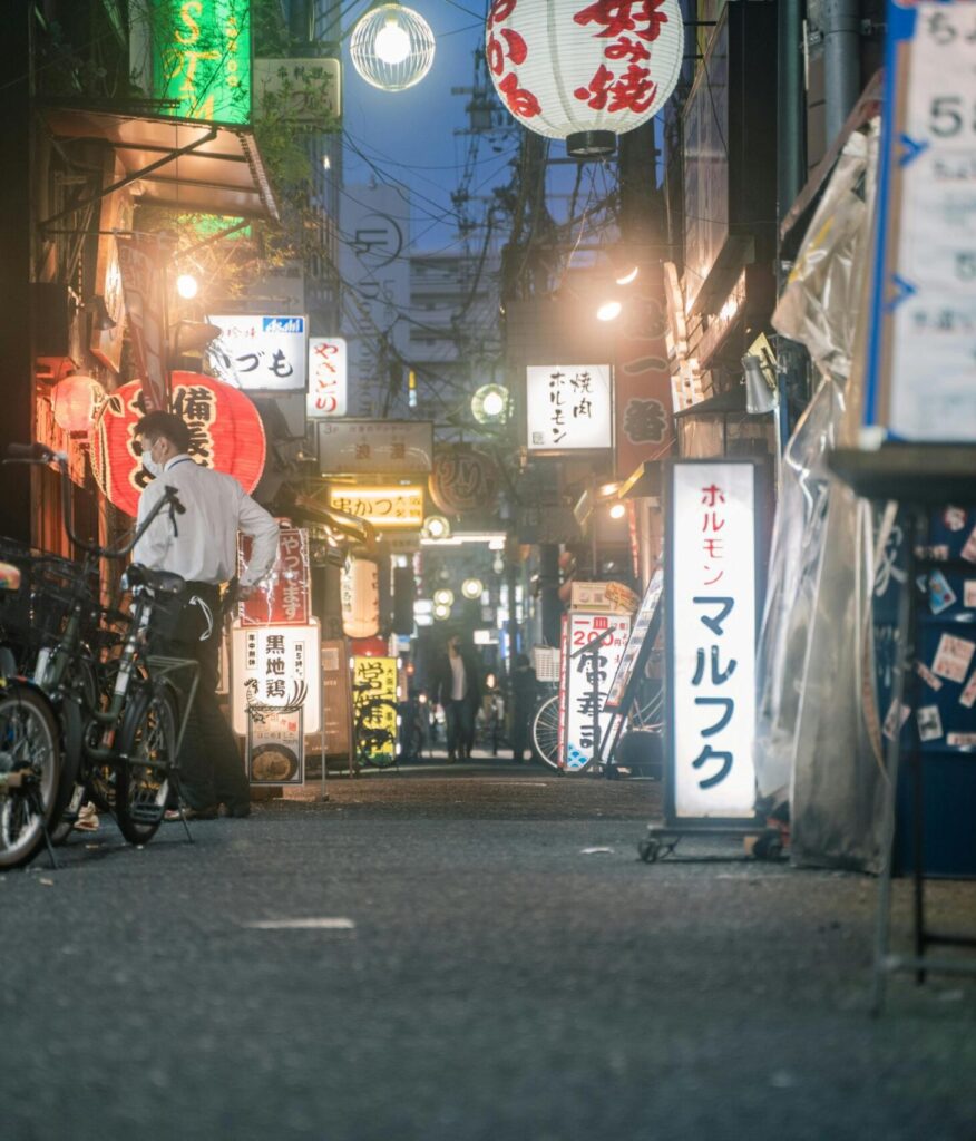 A narrow, dimly lit Japanese alleyway is filled with glowing lanterns, colorful shop signs, and parked bicycles. A man in a white shirt stands near the center of the path, which recedes into a bustling urban background at twilight.