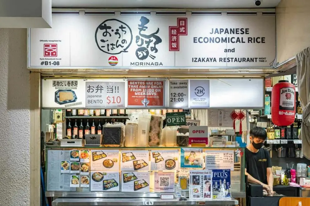 This image shows the storefront of Morinaga, a Japanese Izakaya restaurant that serves "economical rice" and bento boxes. A staff member in a black mask stands behind a counter filled with menu displays, takeaway containers, and various bottled beverages.